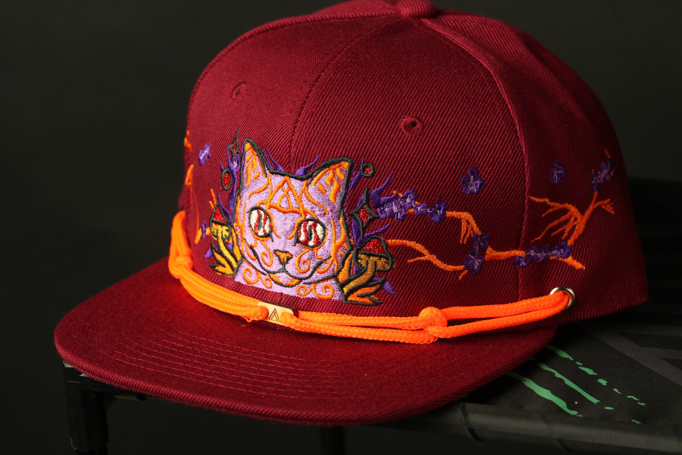 A close-up image of a burgundy baseball cap with a cat design on the front.