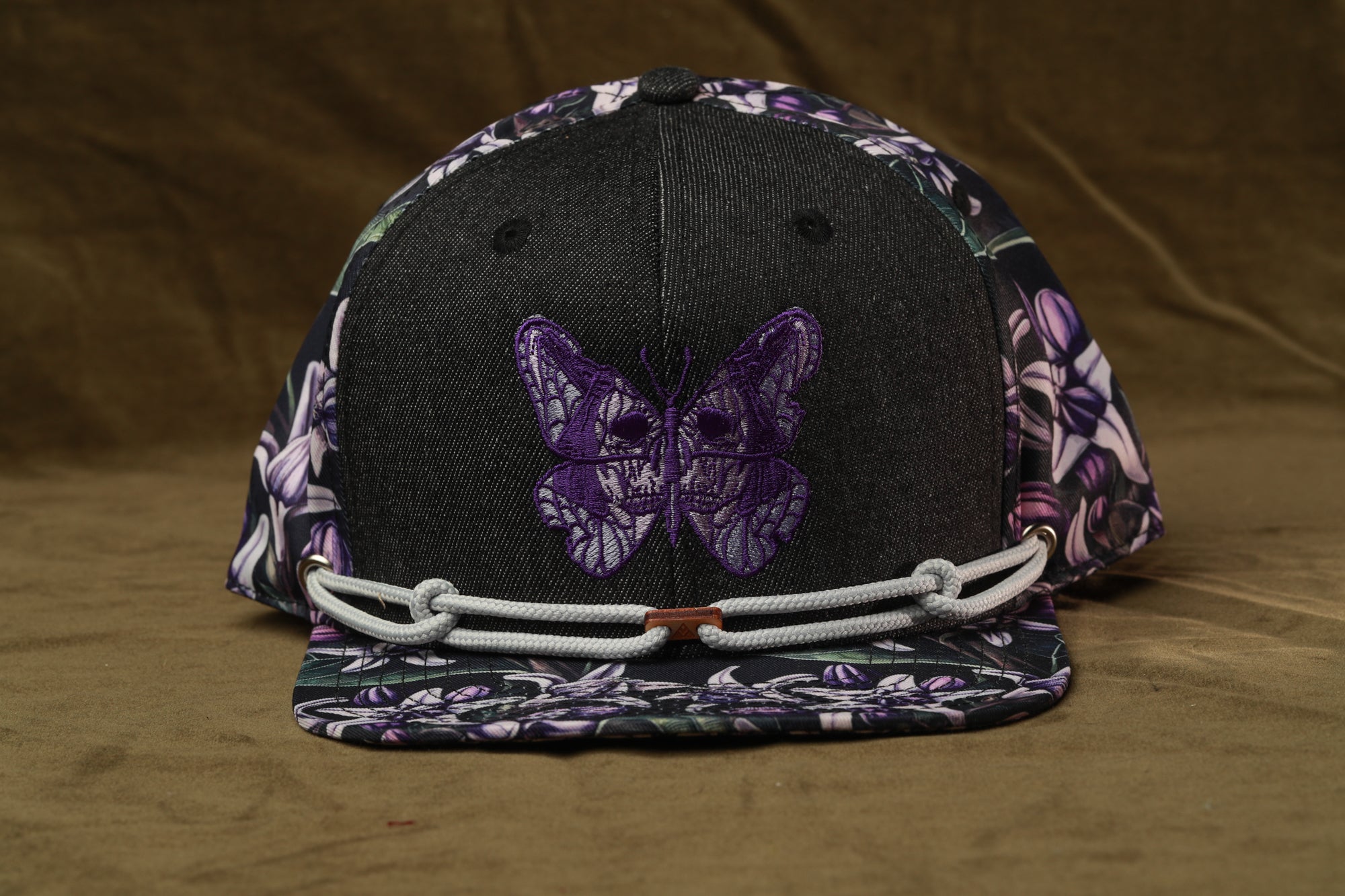 A black baseball cap with a purple butterfly embroidered on it, featuring a floral pattern on the brim and a white rope strap.