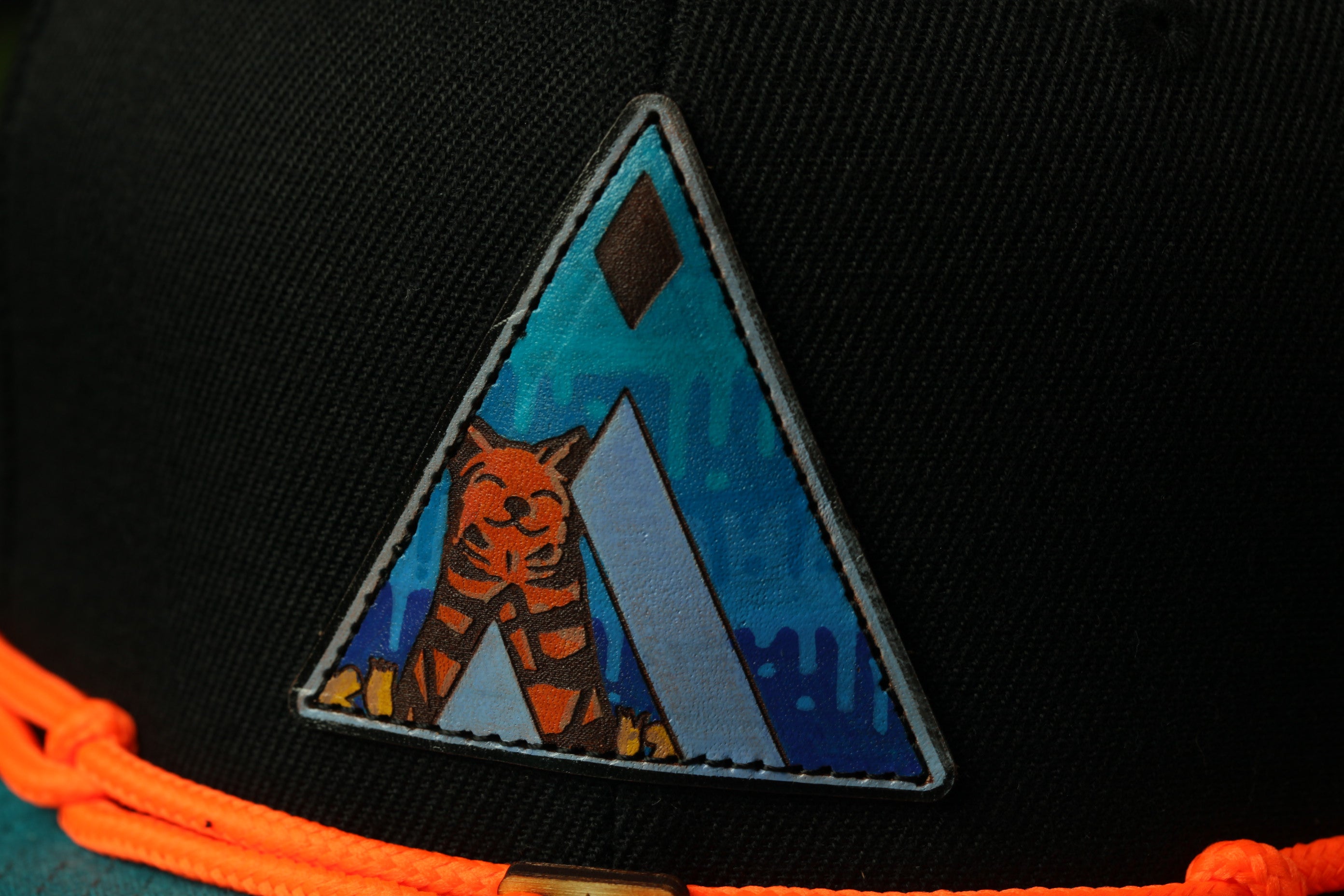 A close-up view of a black baseball cap with a colorful triangular patch featuring an illustration of a cat.