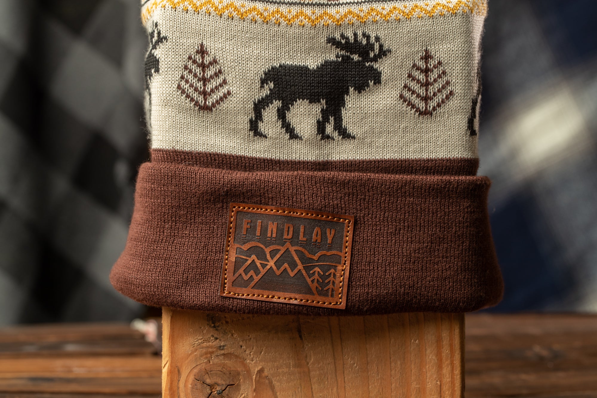 A close-up image of a brown and white knitted beanie with a moose design, placed on a wooden surface.