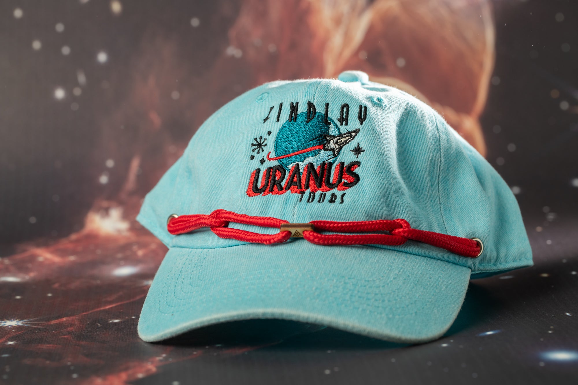 A light blue baseball cap with a red string and the text "URANUS" printed on it, set against a backdrop of a space-themed image.