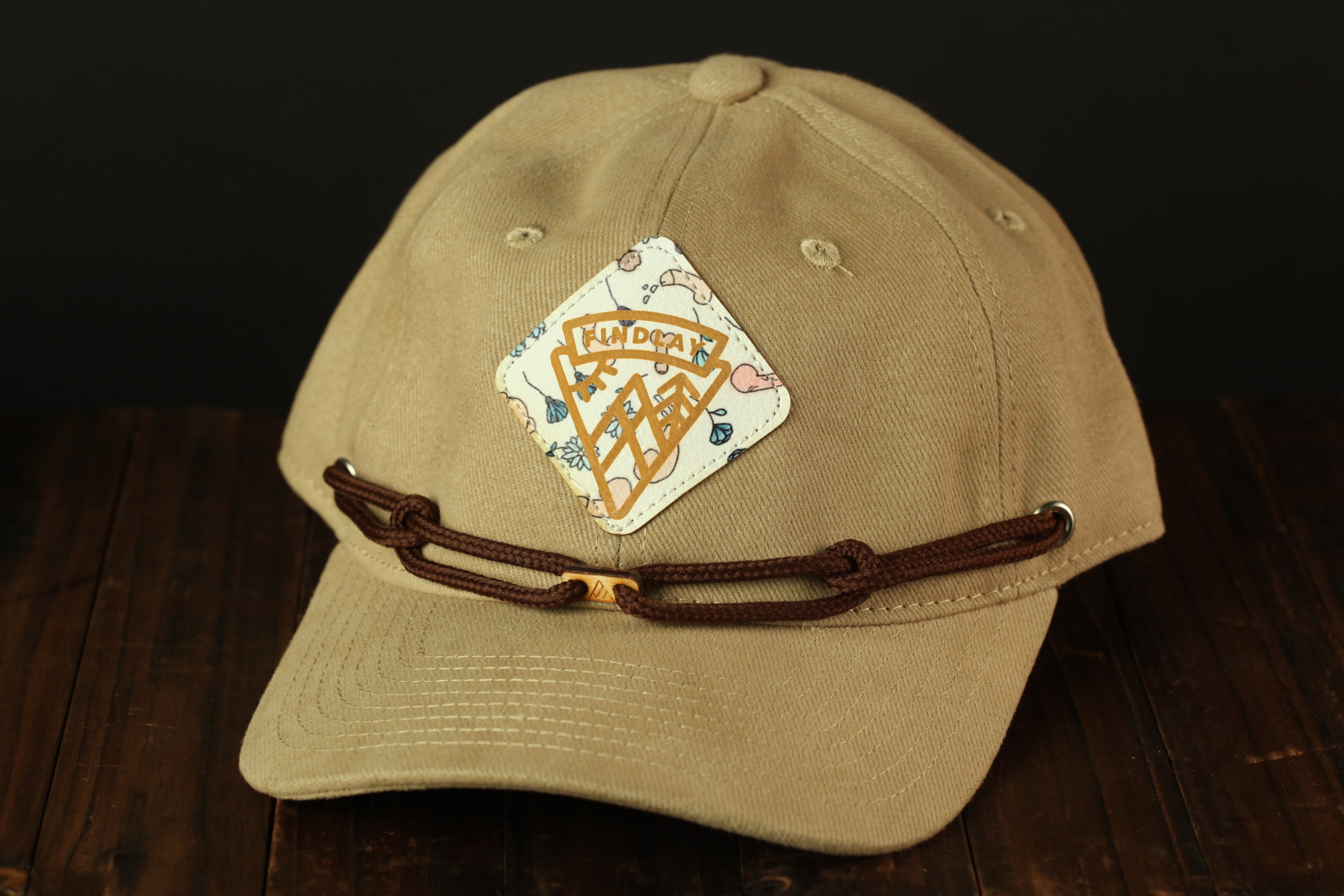 A beige baseball cap with a decorative patch and brown leather strap is displayed on a wooden surface.