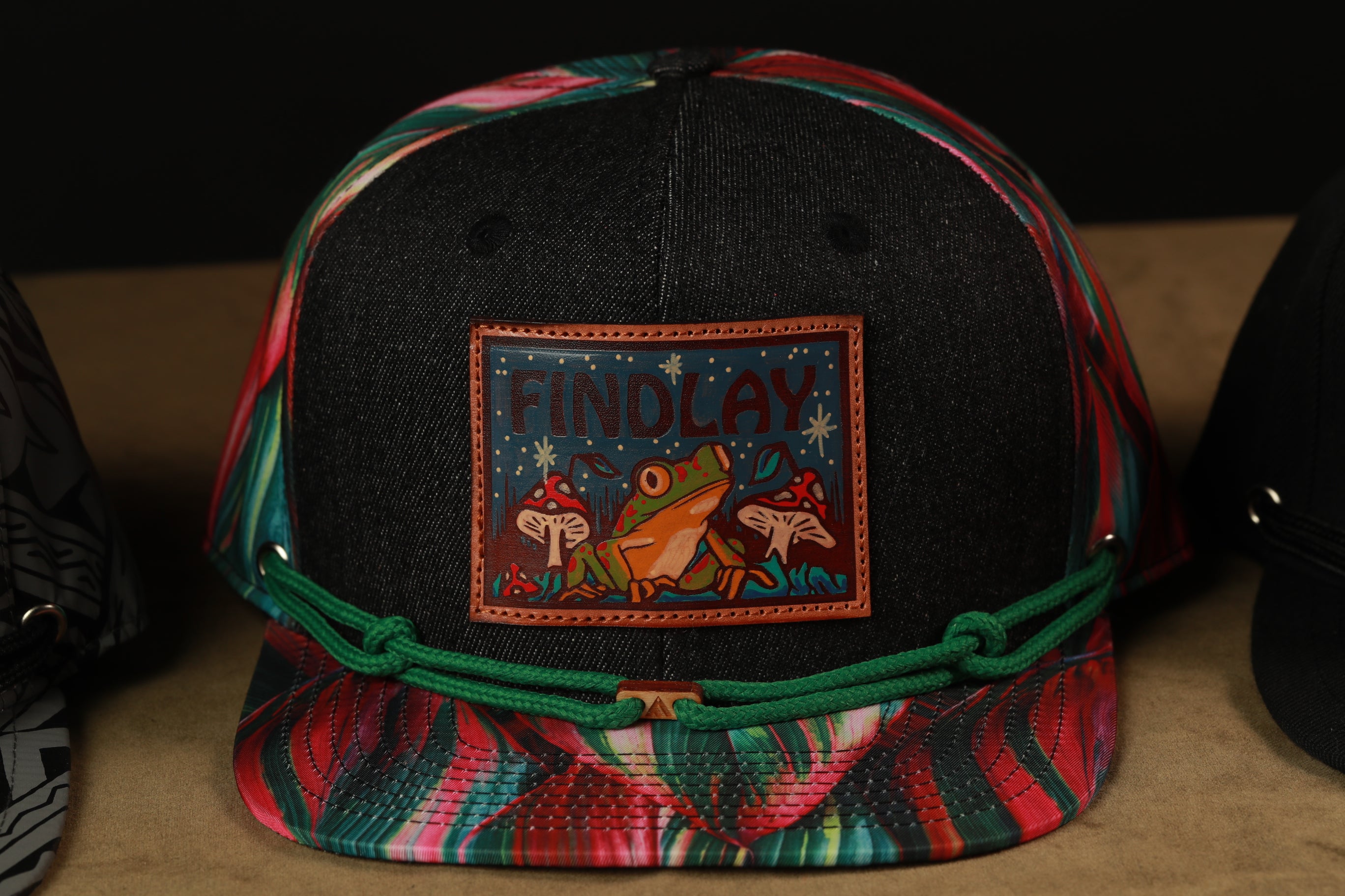 A colorful baseball cap with a patch featuring a frog and mushrooms, along with the text "FIND LAY".