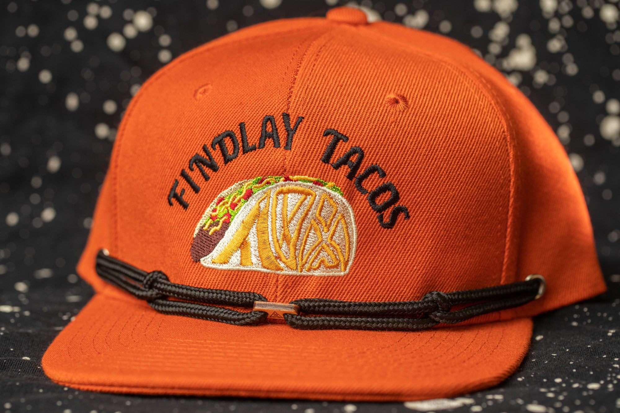 An orange baseball cap with the text "Findlay Tacos" embroidered on it, featuring a graphic of a taco.