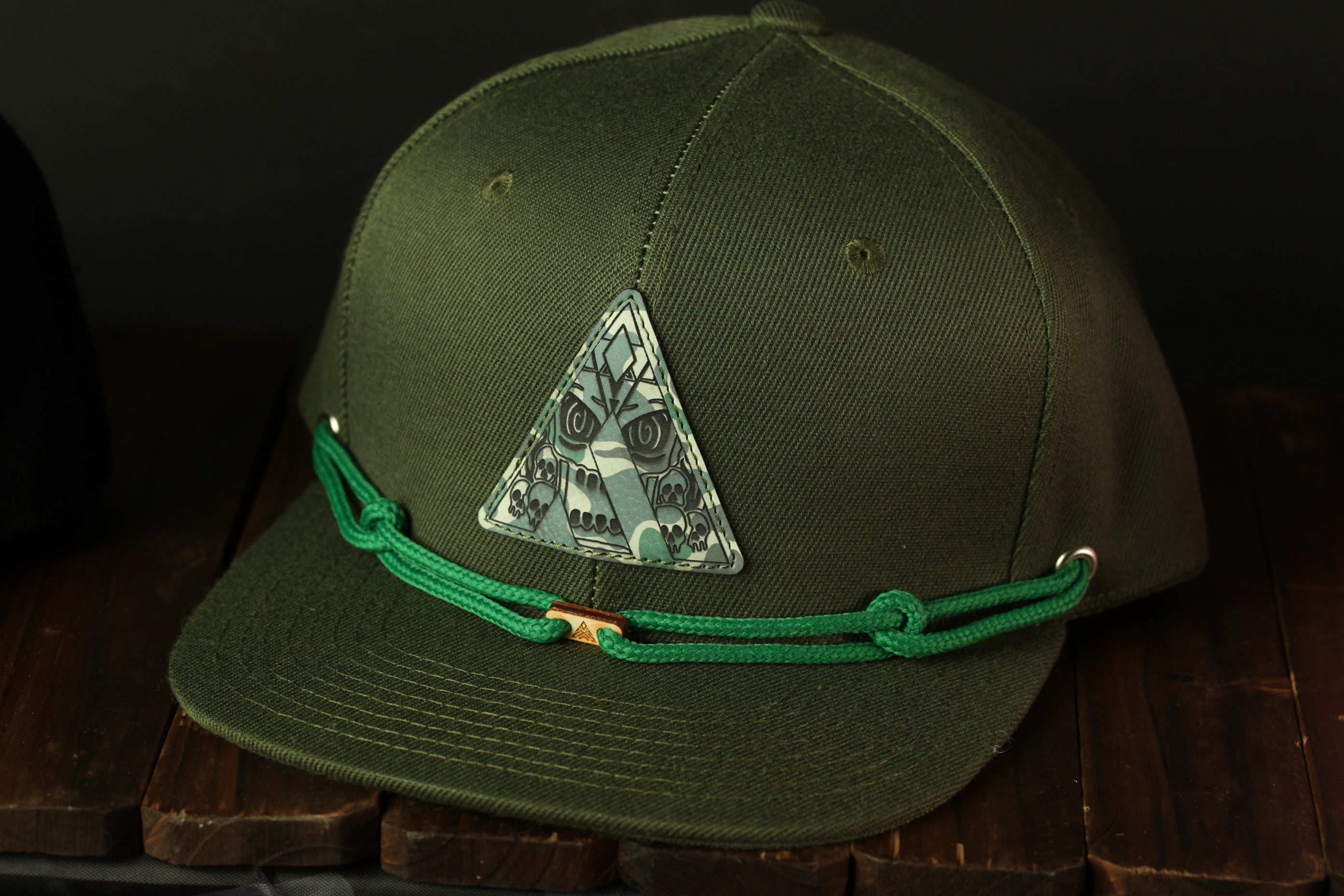 A green baseball cap with a triangular patch on the front, featuring a skull and crossbones design, is displayed on a wooden surface.