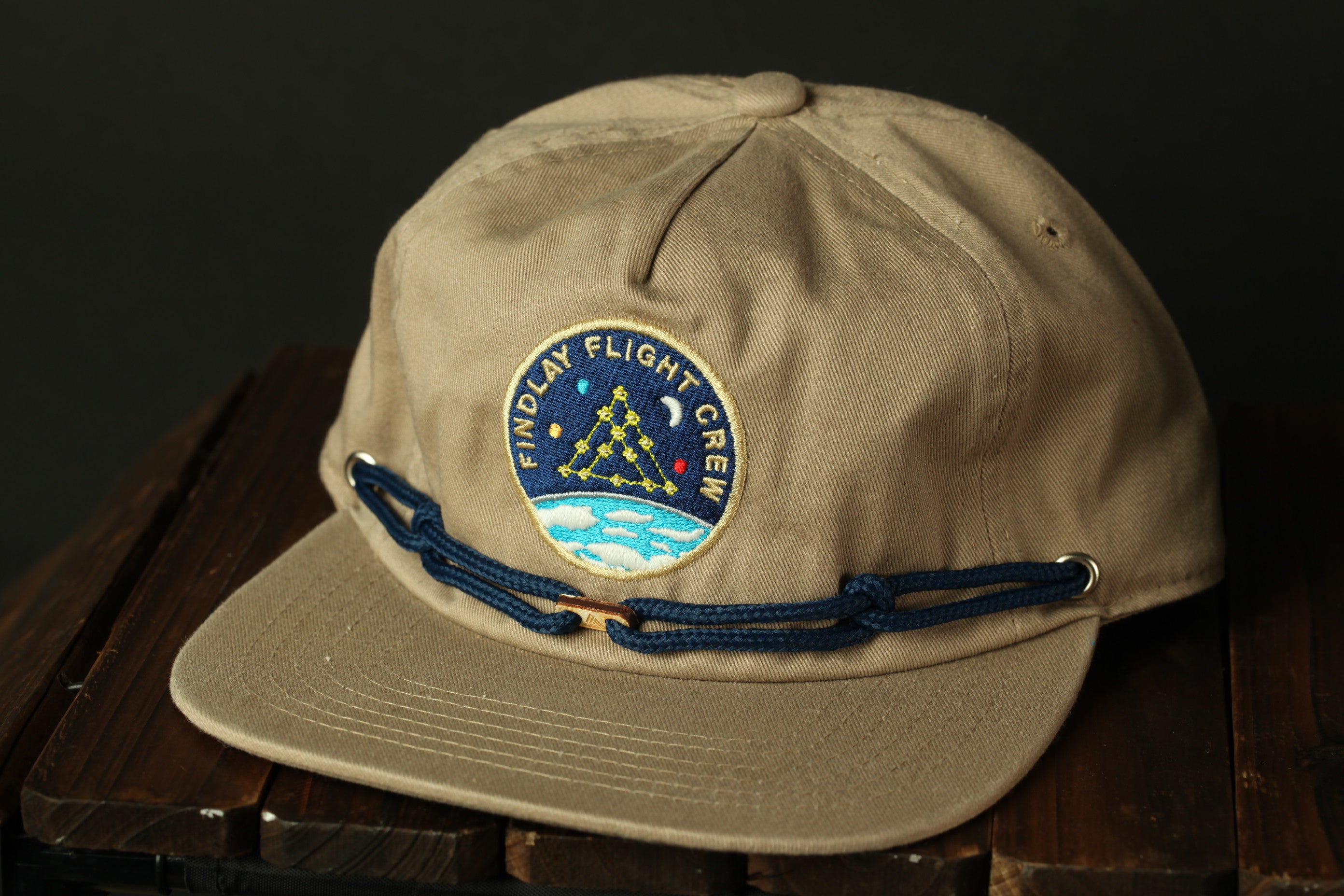 A beige baseball cap with a blue band and a circular patch on the front, featuring a design with stars and a blue background.