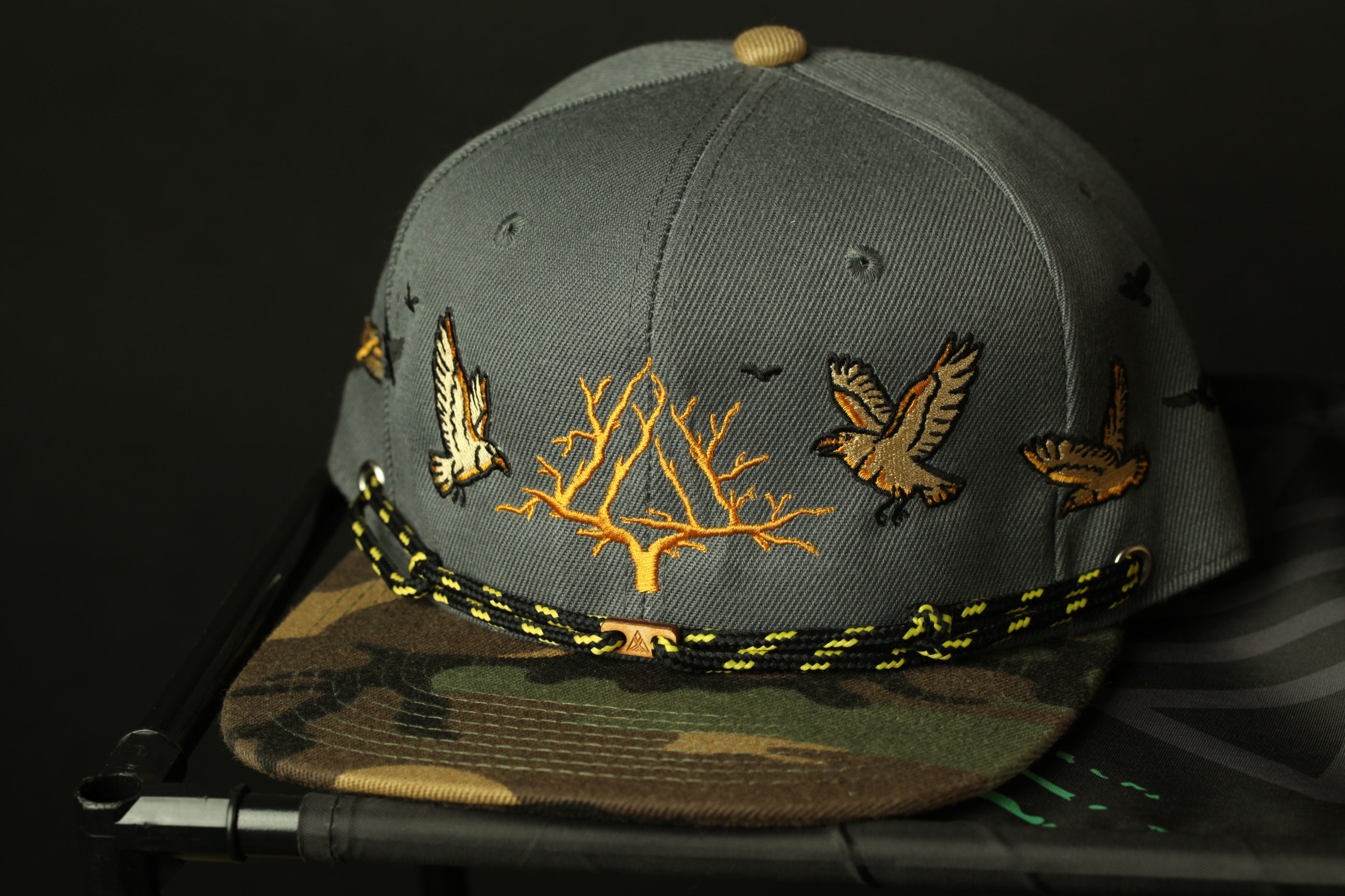A gray baseball cap with a camouflage pattern and embroidered designs of birds on the brim.