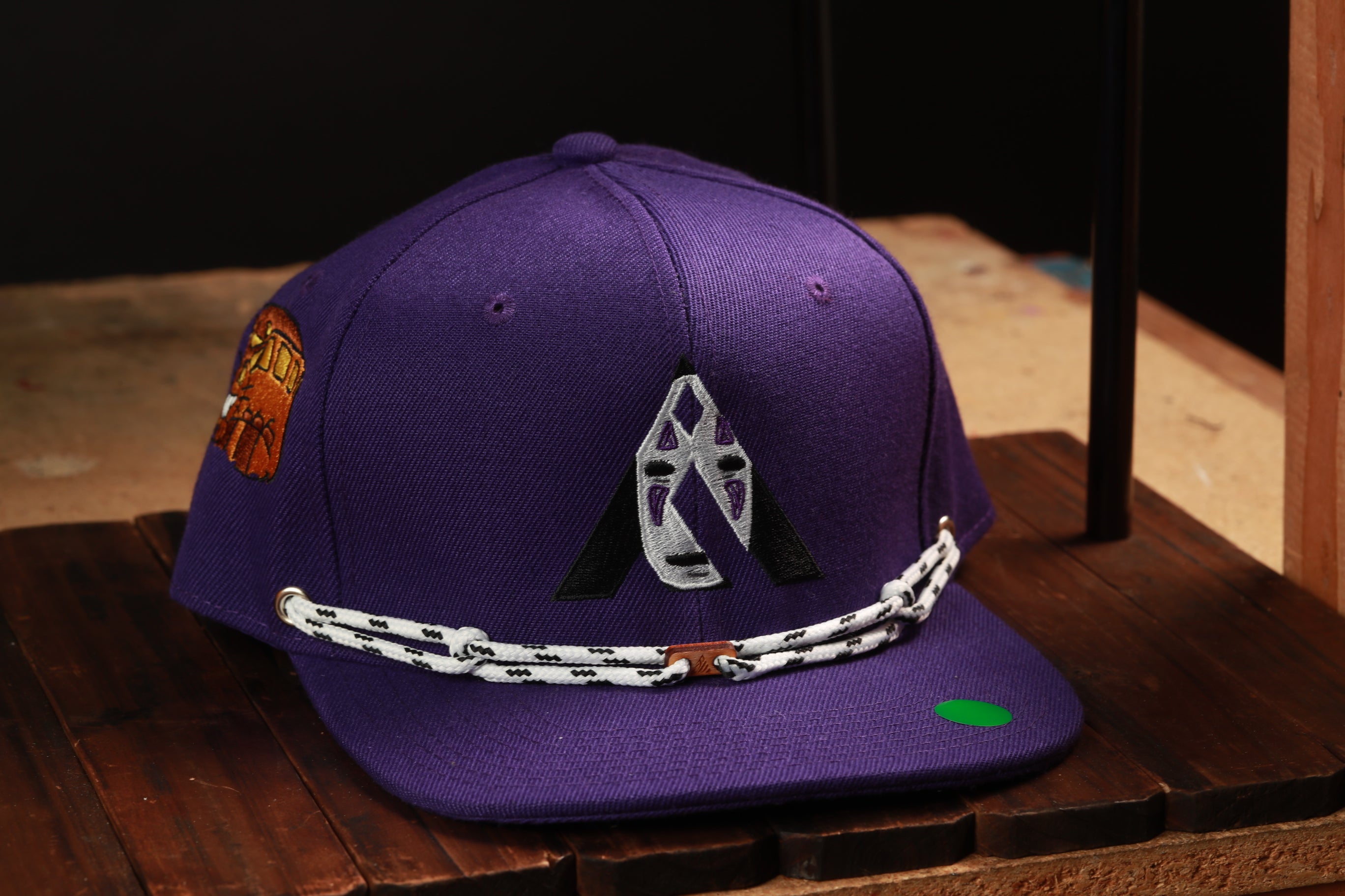 A purple baseball cap with a white and black chain strap, featuring a logo on the front, is displayed on a wooden surface.