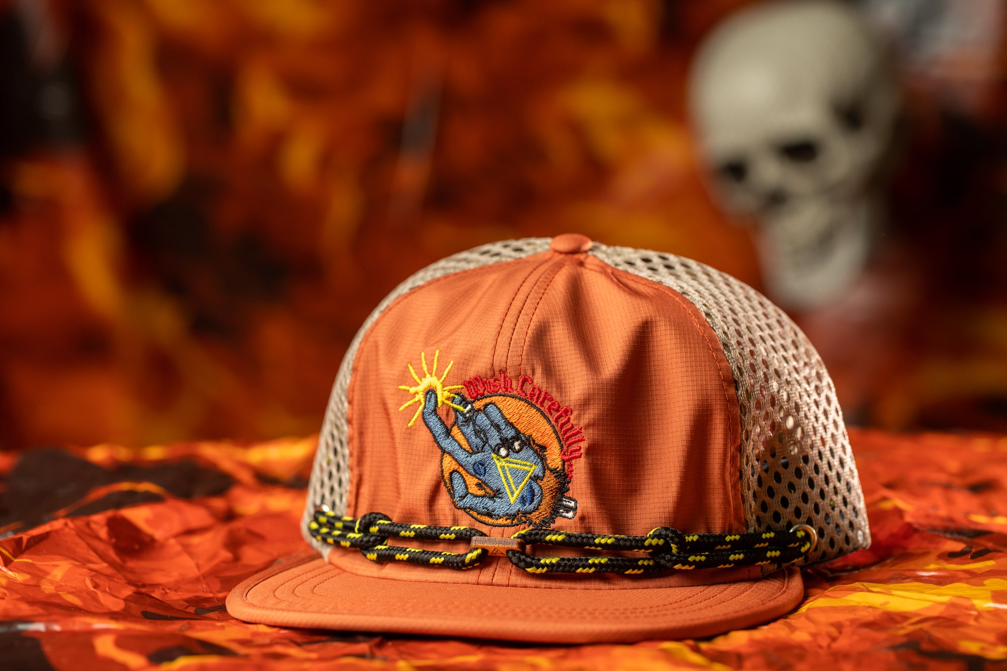 An orange baseball cap with a mesh back and a graphic design on the front, placed on a surface with a blurred background that includes a skull and some orange leaves.