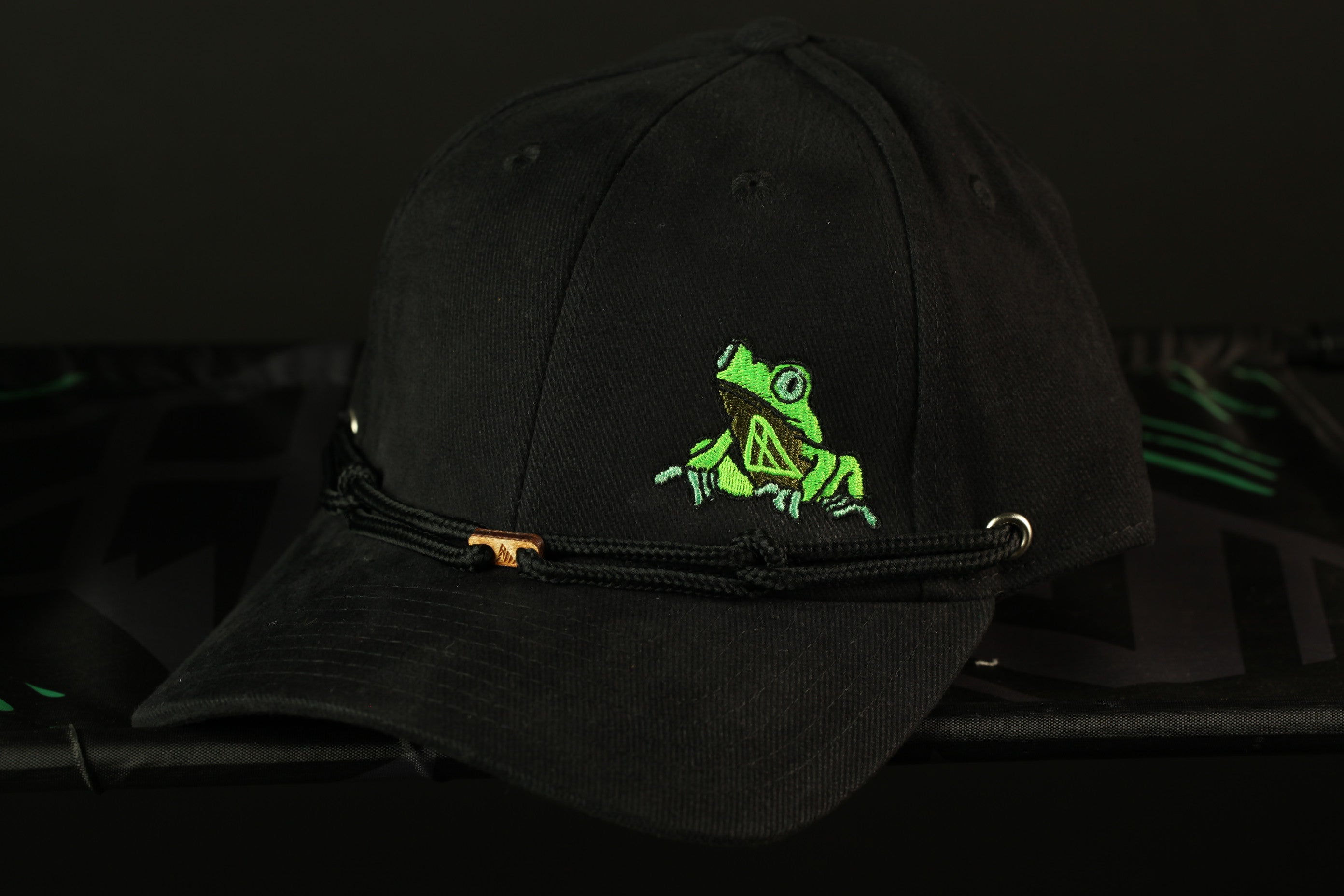 A black baseball cap with a green frog embroidered on it, resting on a black surface.