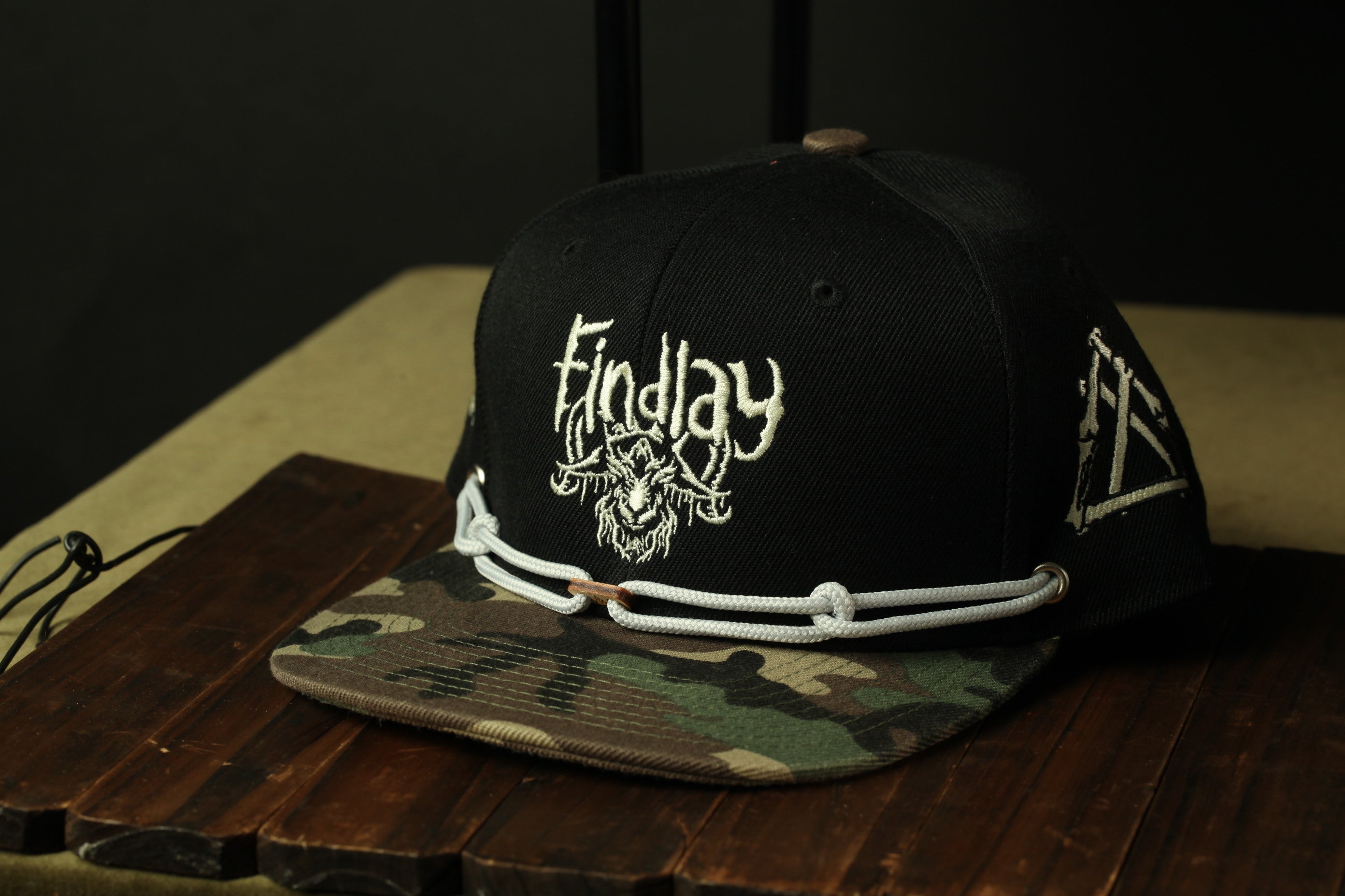 A black baseball cap with a camouflage pattern and the word "Findlay" printed on it, sitting on a wooden surface.