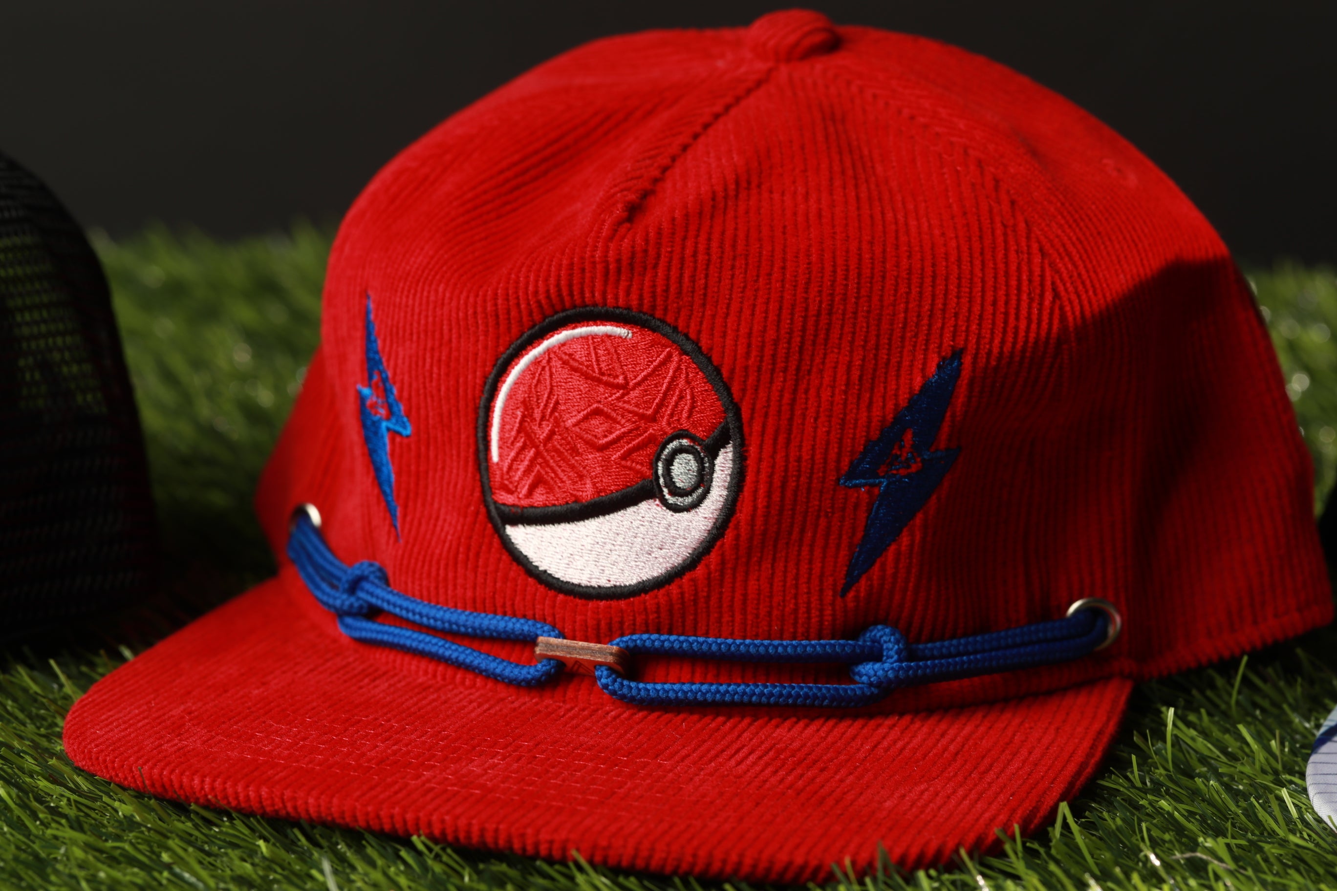 A red baseball cap with a Pokemon-themed design, including a red ball and blue lightning bolts, is shown on a grassy surface.