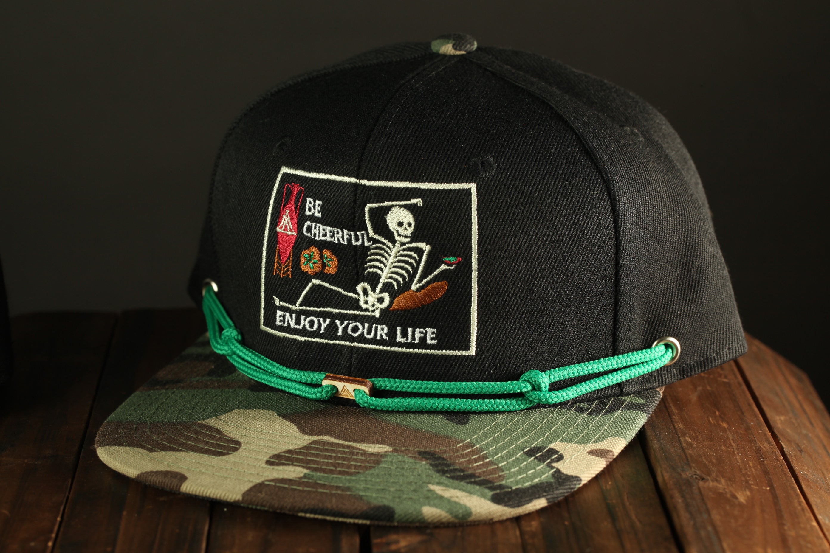 A black baseball cap with a camouflage pattern and a patch on the front that says "Be cheerful, enjoy your life".