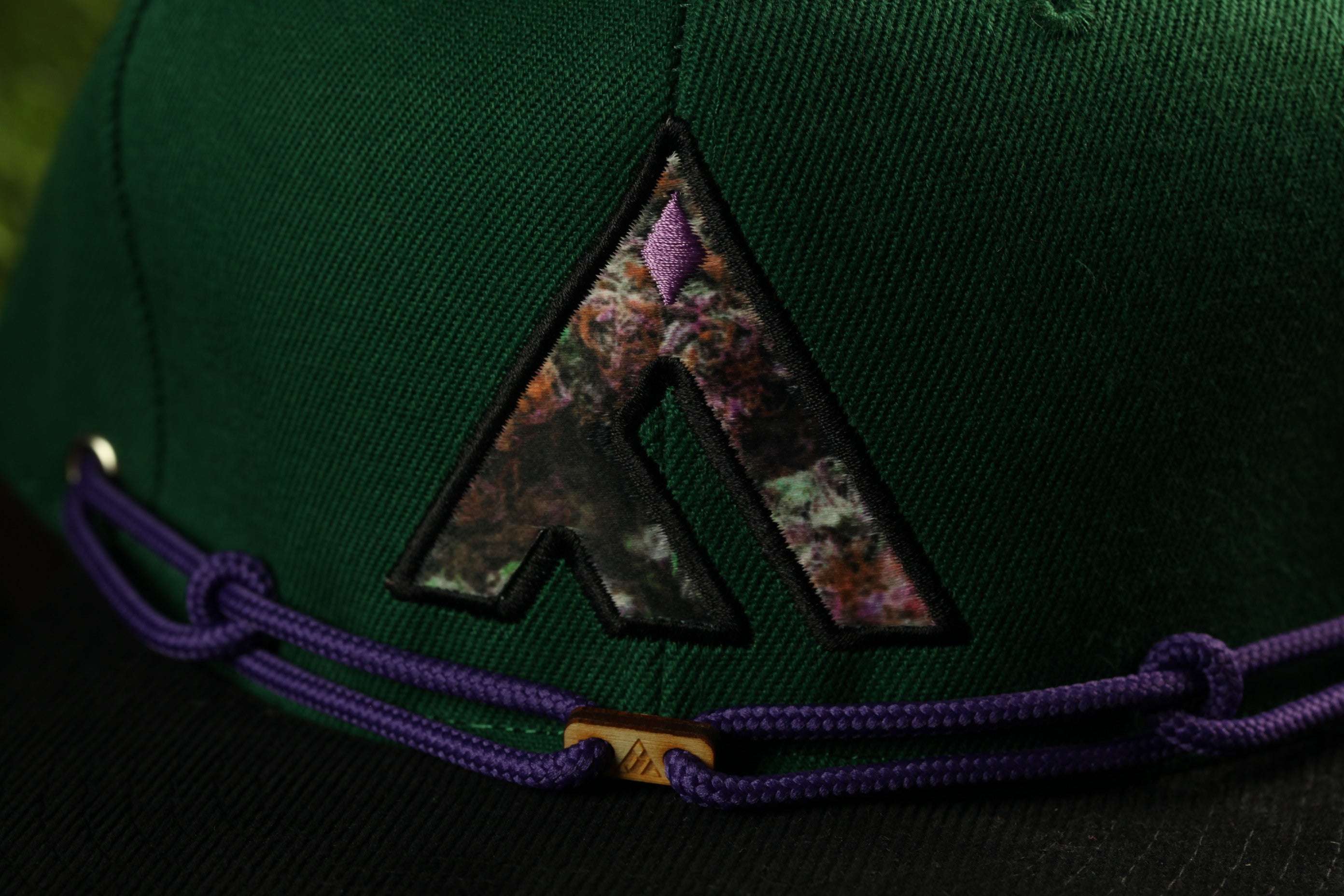 A close-up view of a green baseball cap with a purple and black embroidered logo on the front.