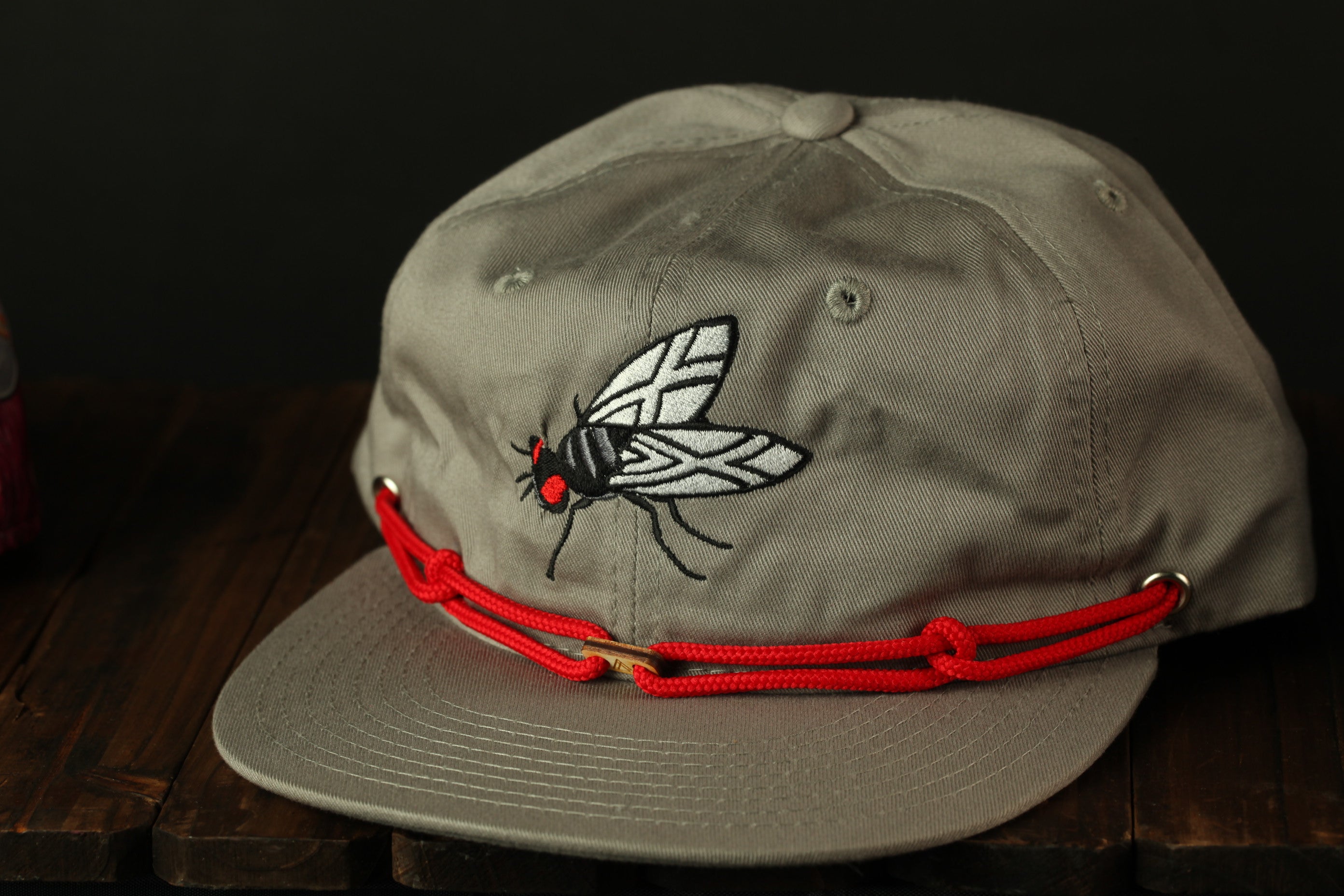 A gray baseball cap with a red string and a black and white fly embroidered on the front.