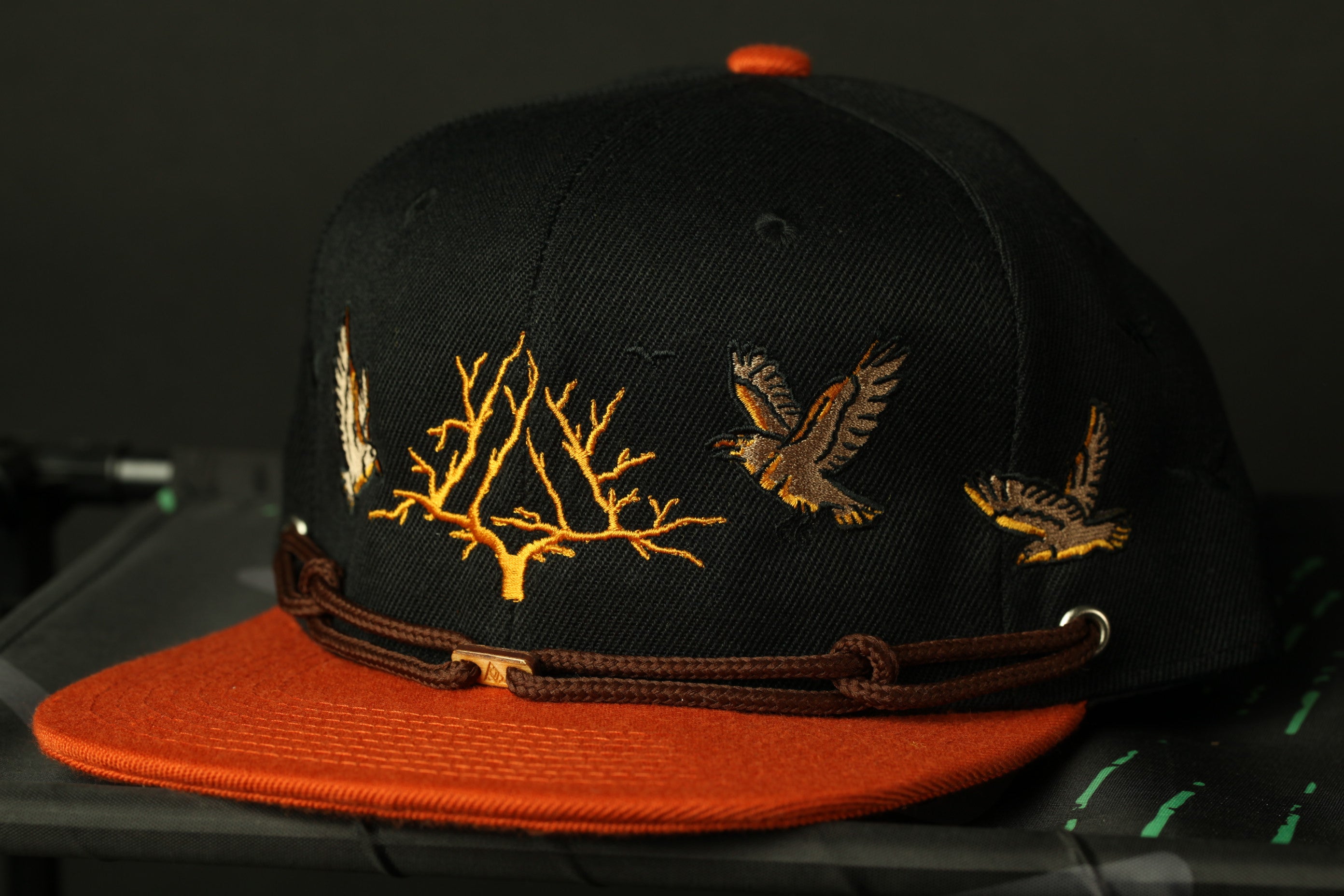 A black baseball cap with orange trim and gold embroidered designs, including a tree and two birds, is displayed against a dark background.