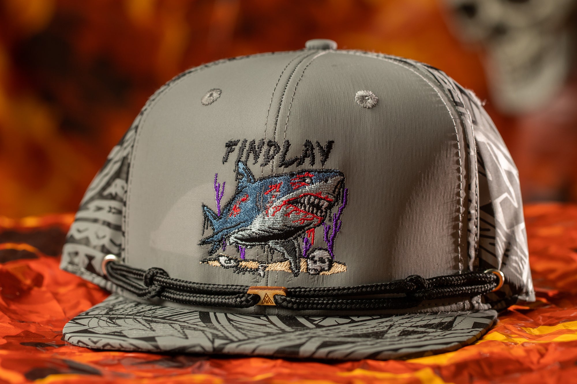 A gray baseball cap with a graphic of a shark and the word "FINDPLAY" printed on it, placed on a red and orange background.