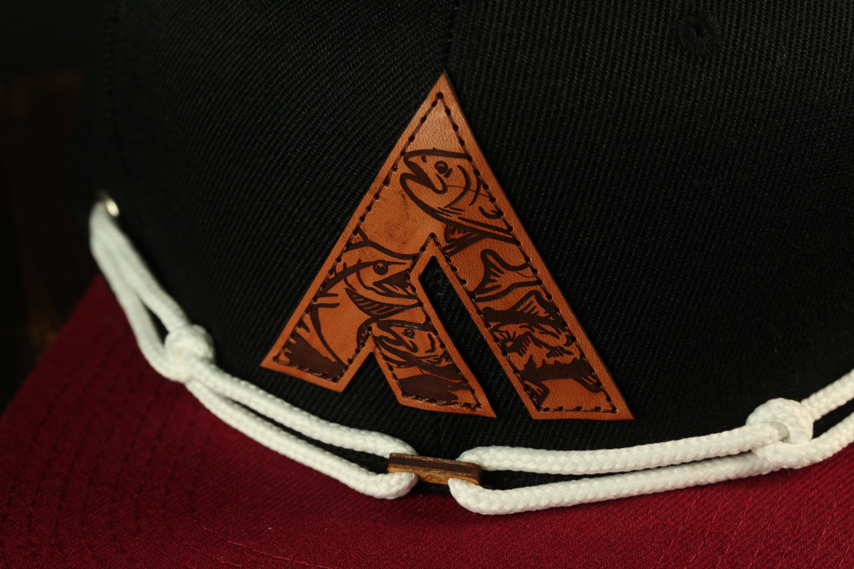A close-up view of a black baseball cap with a white rope and a leather-like logo on the front.