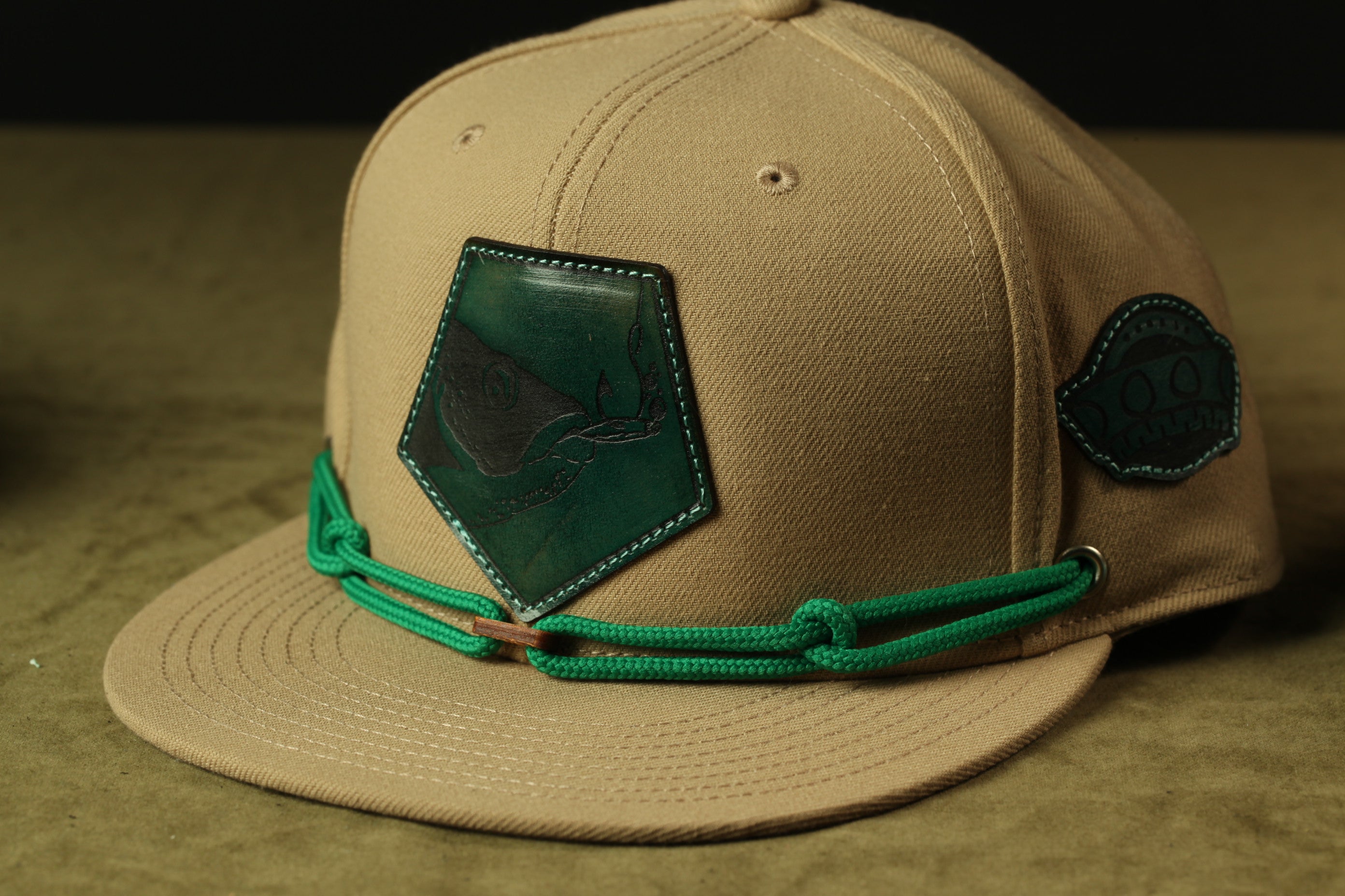 A beige baseball cap with a green and black patch on the front and a green string around the brim.