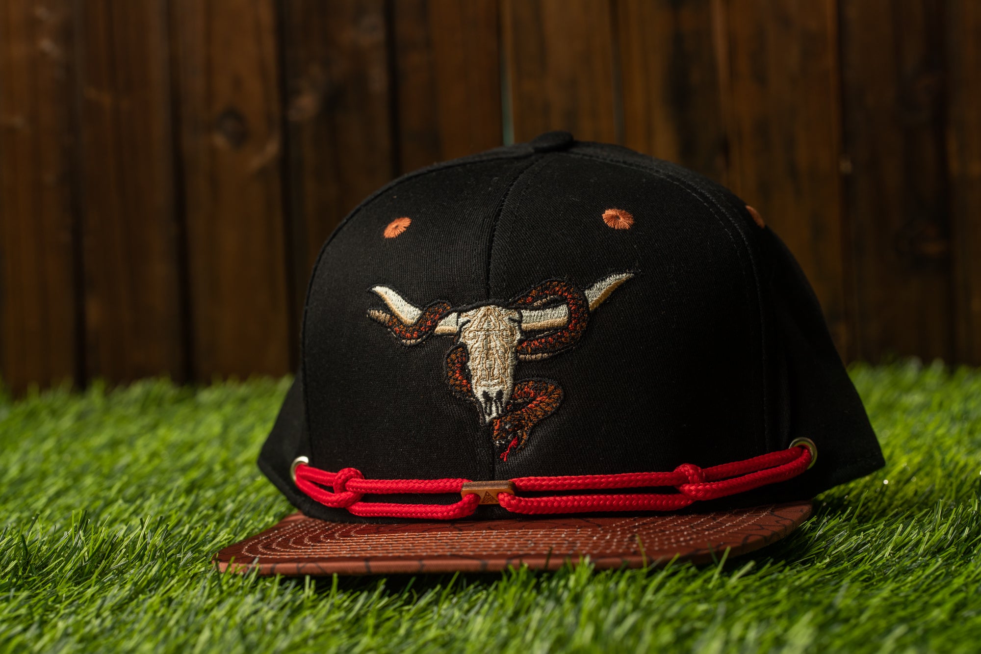 A black baseball cap with a red strap and a graphic of a bull's head is placed on a grassy surface.
