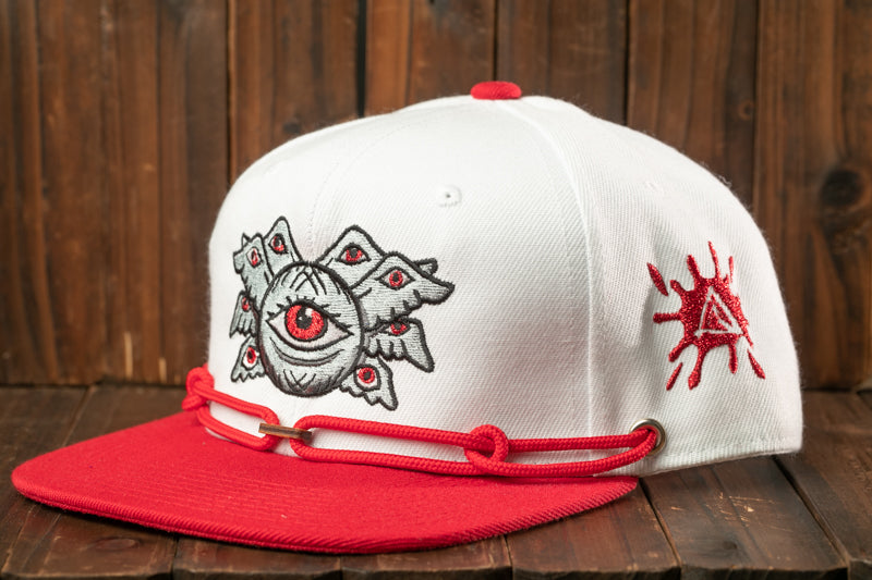A white baseball cap with a red brim and a unique eye design on the front, placed on a wooden surface.
