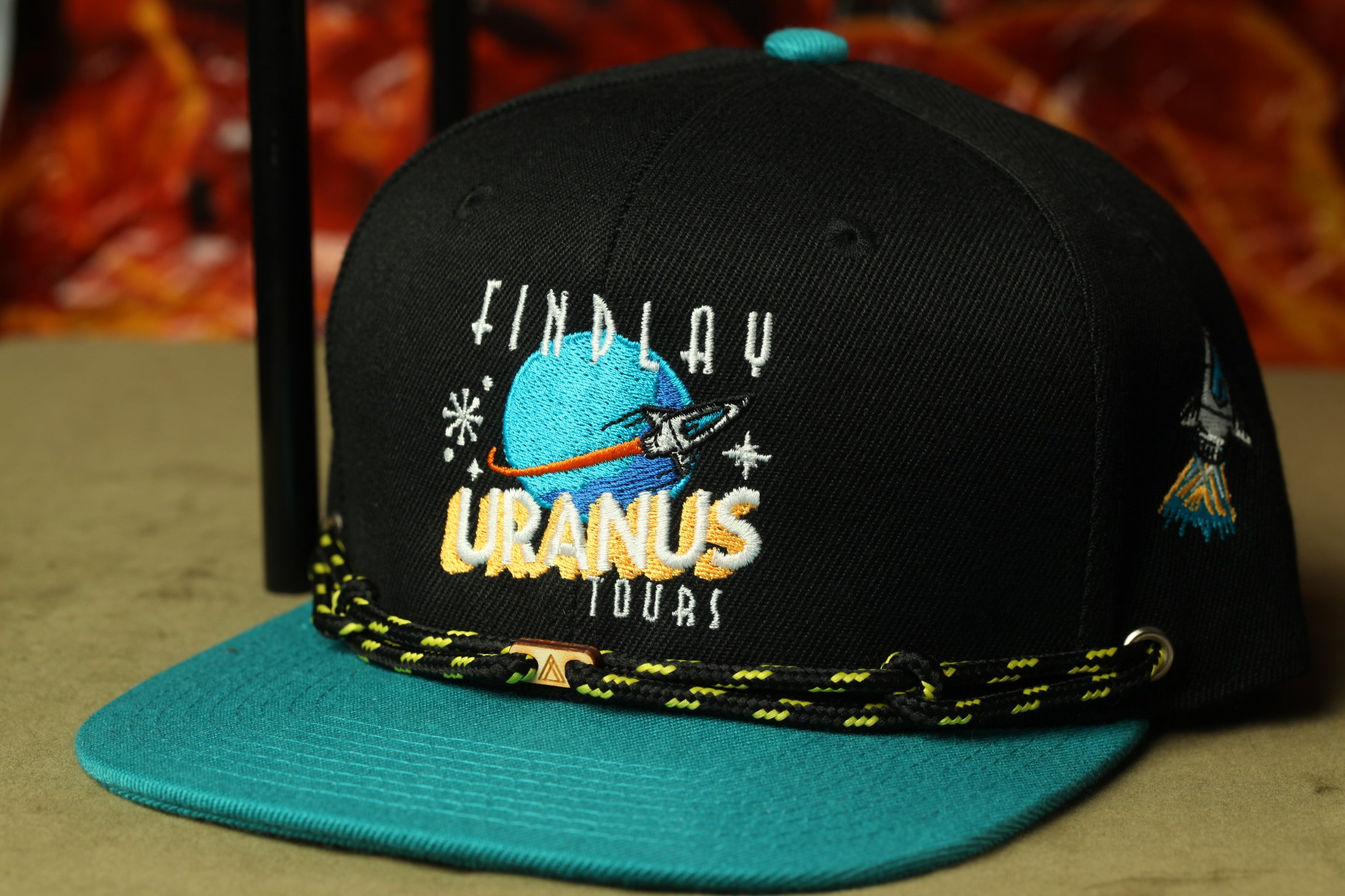 A black baseball cap with a teal brim and a black and yellow braided strap. The cap has a logo on the front that says "FINDLAY URANUS TOUR".