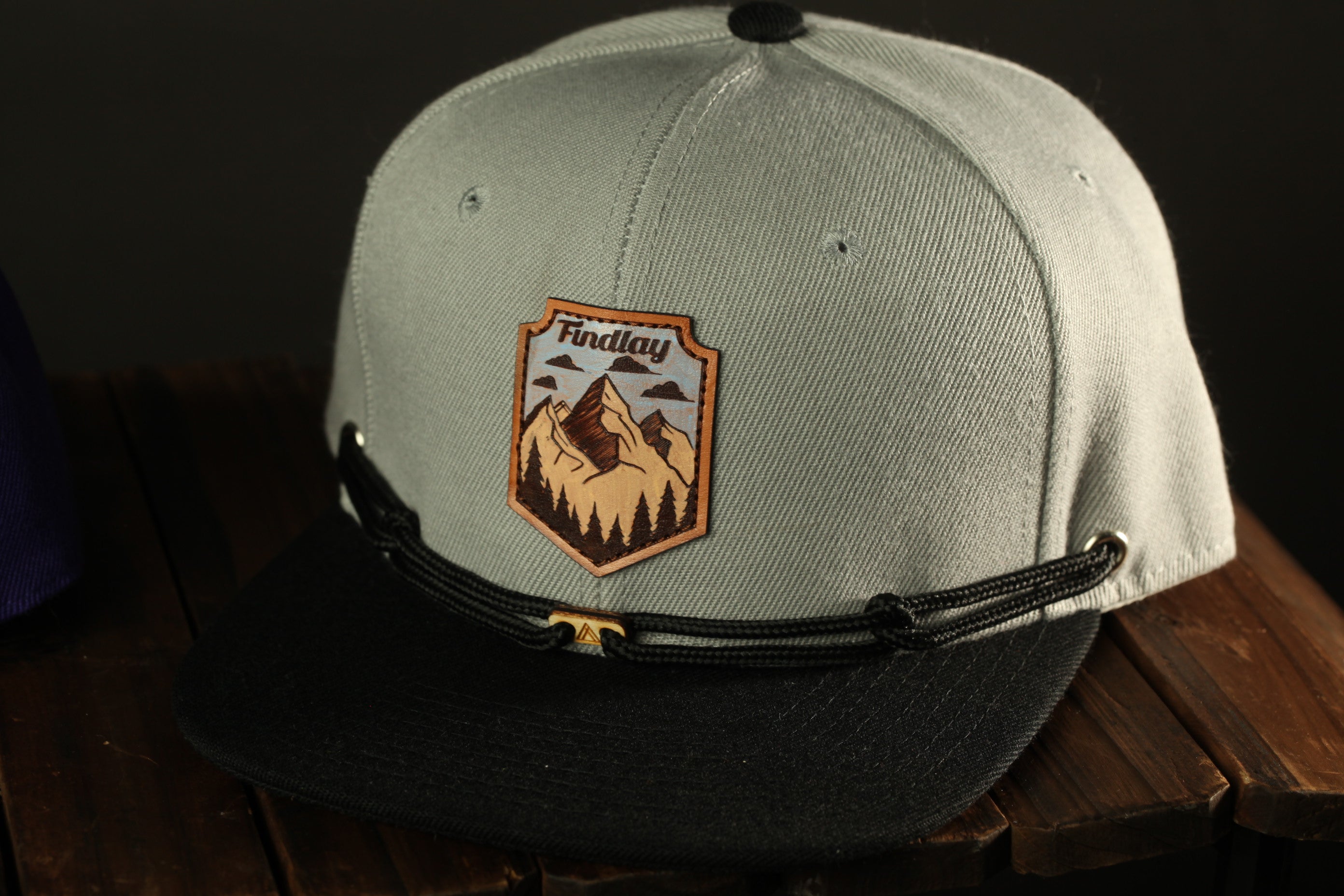A gray baseball cap with a black brim and a black and white mountain patch on the front, sitting on a wooden surface.