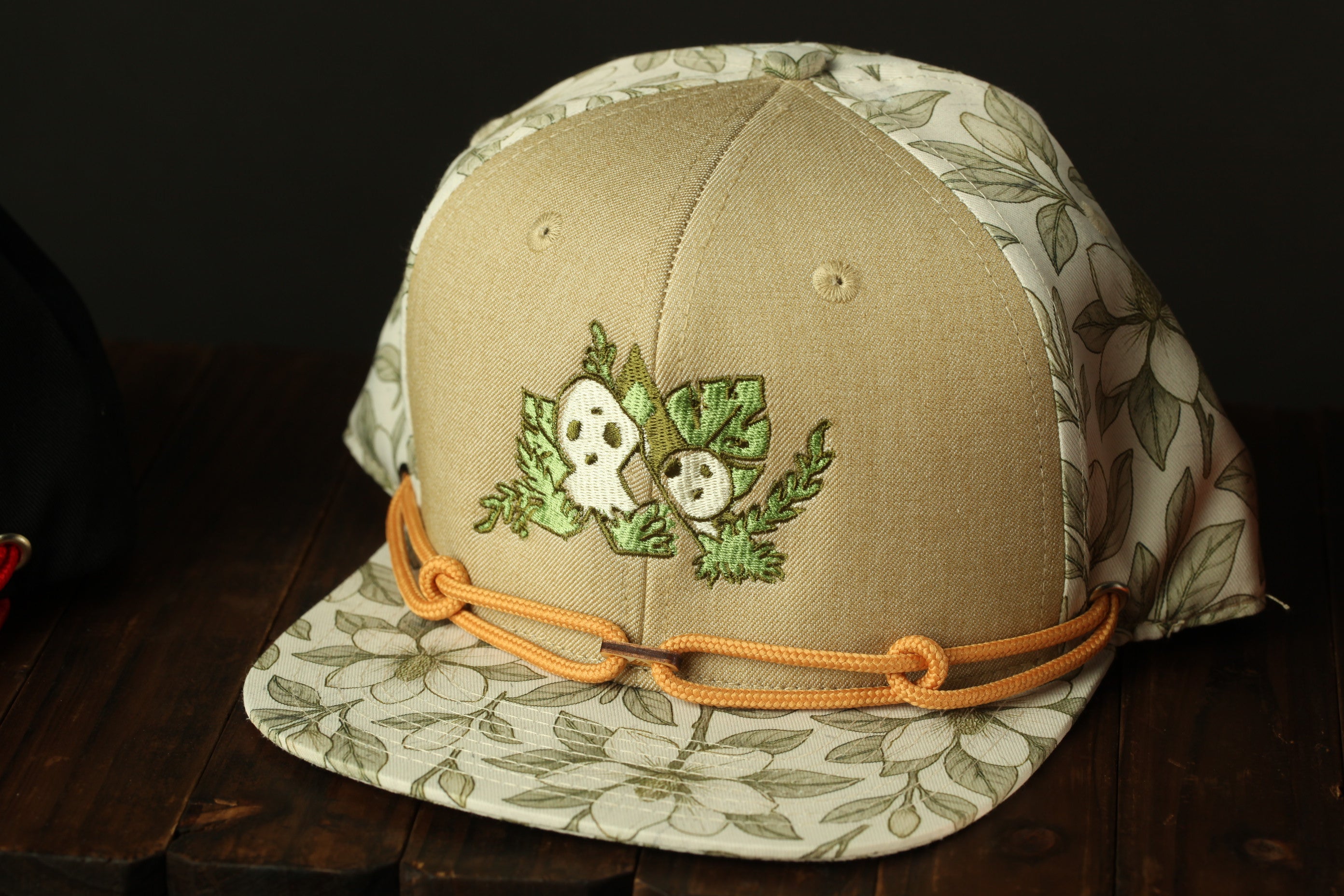 A beige baseball cap with a floral pattern and a small embroidered design on the front, sitting on a wooden surface.
