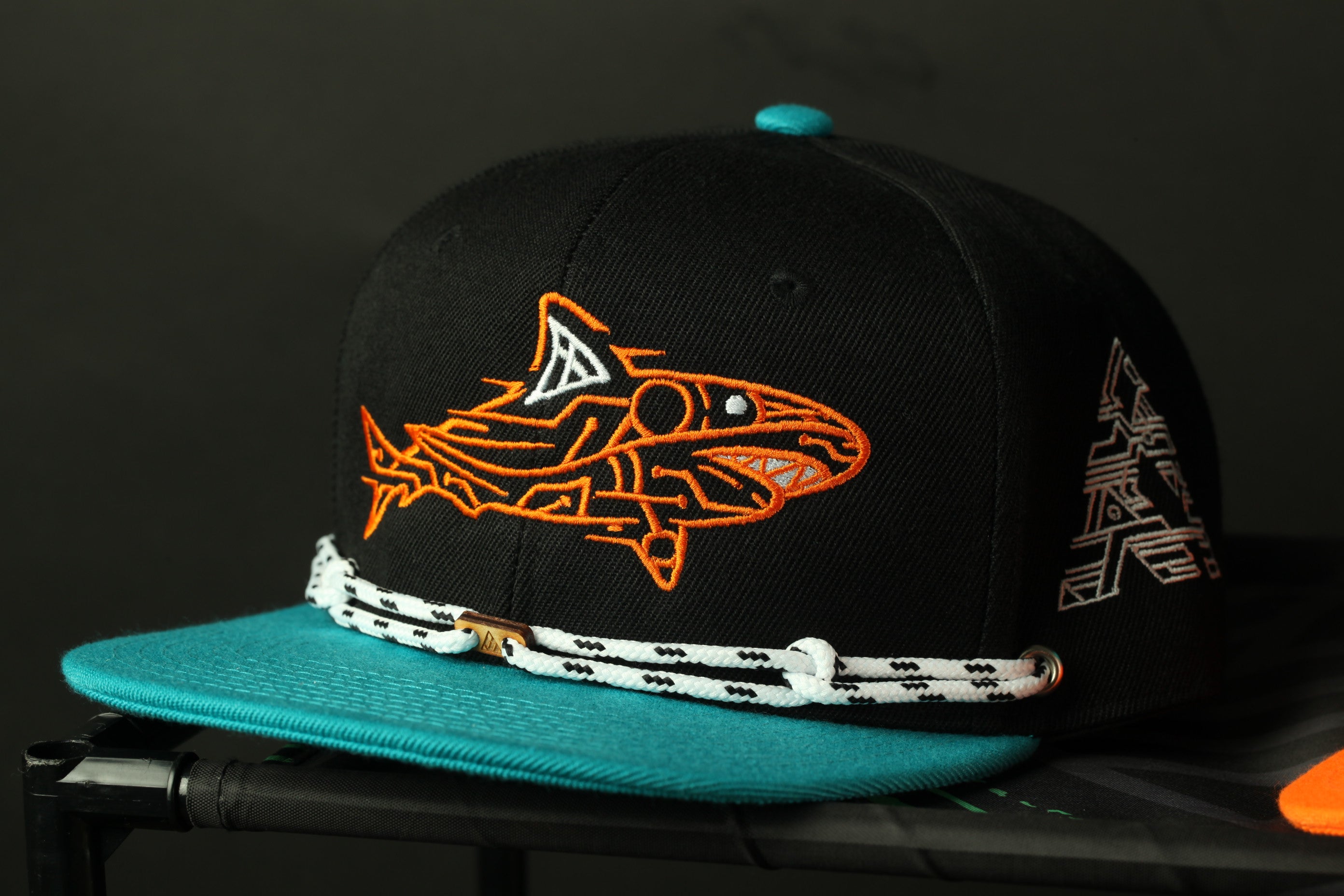 A black baseball cap with an orange and black shark design, featuring a teal brim and a white lace strap.