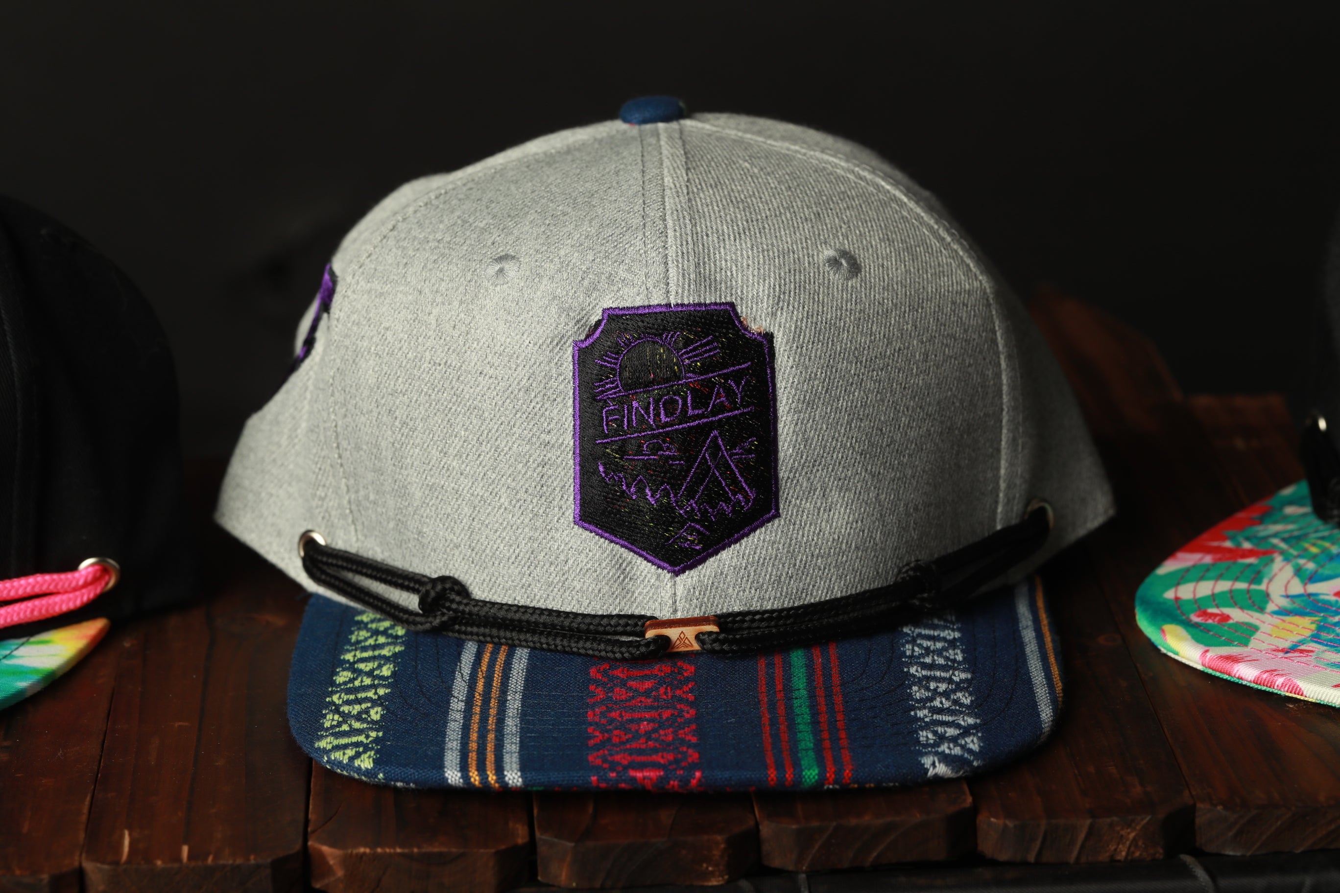 A gray baseball cap with a purple and black patch on the front, featuring a graphic design, is displayed on a wooden surface.