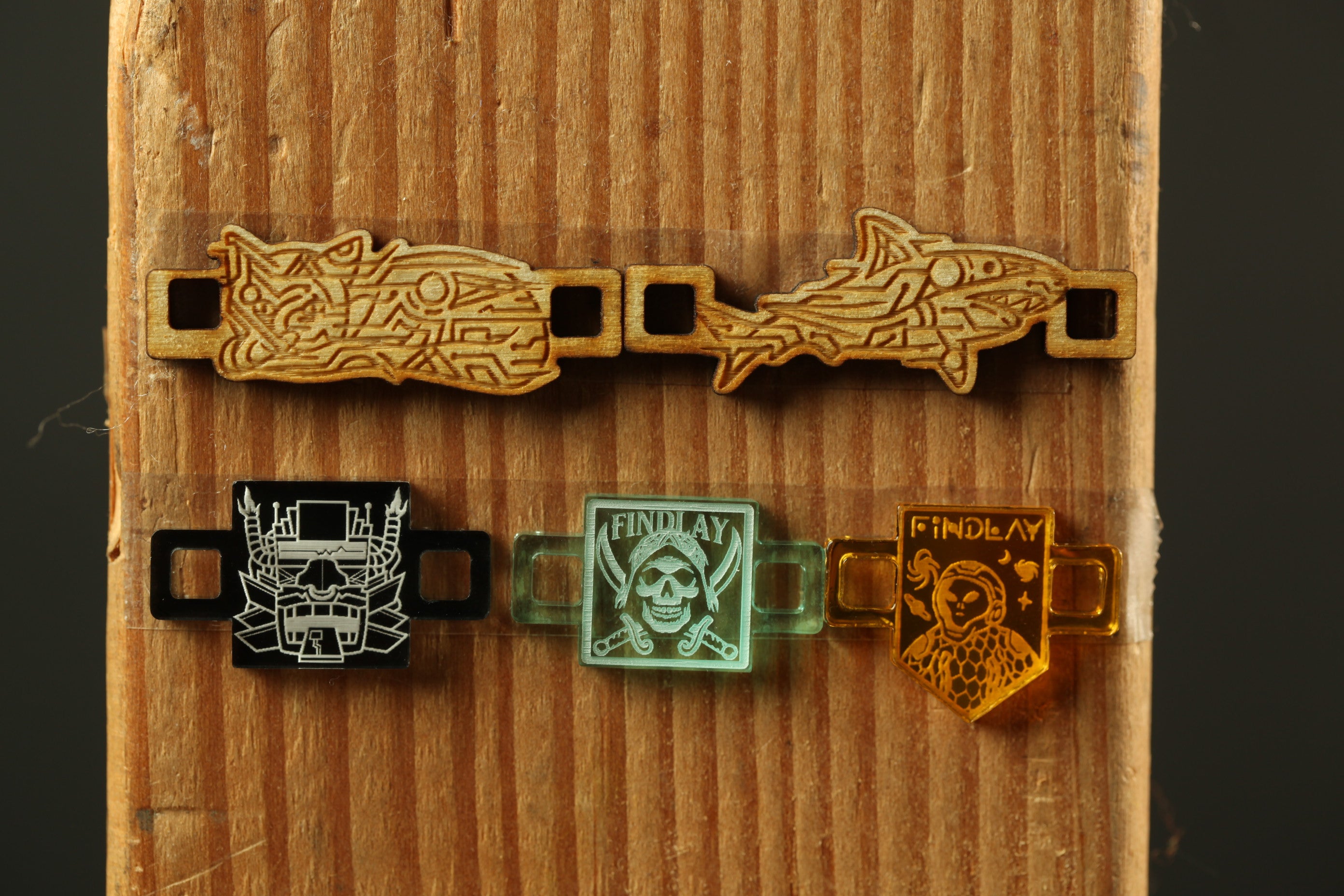 a wooden surface with various objects attached to it, including a pair of wooden shark-shaped charms, a black and white skull-shaped charm, and a green and orange charm with a pirate-like design.