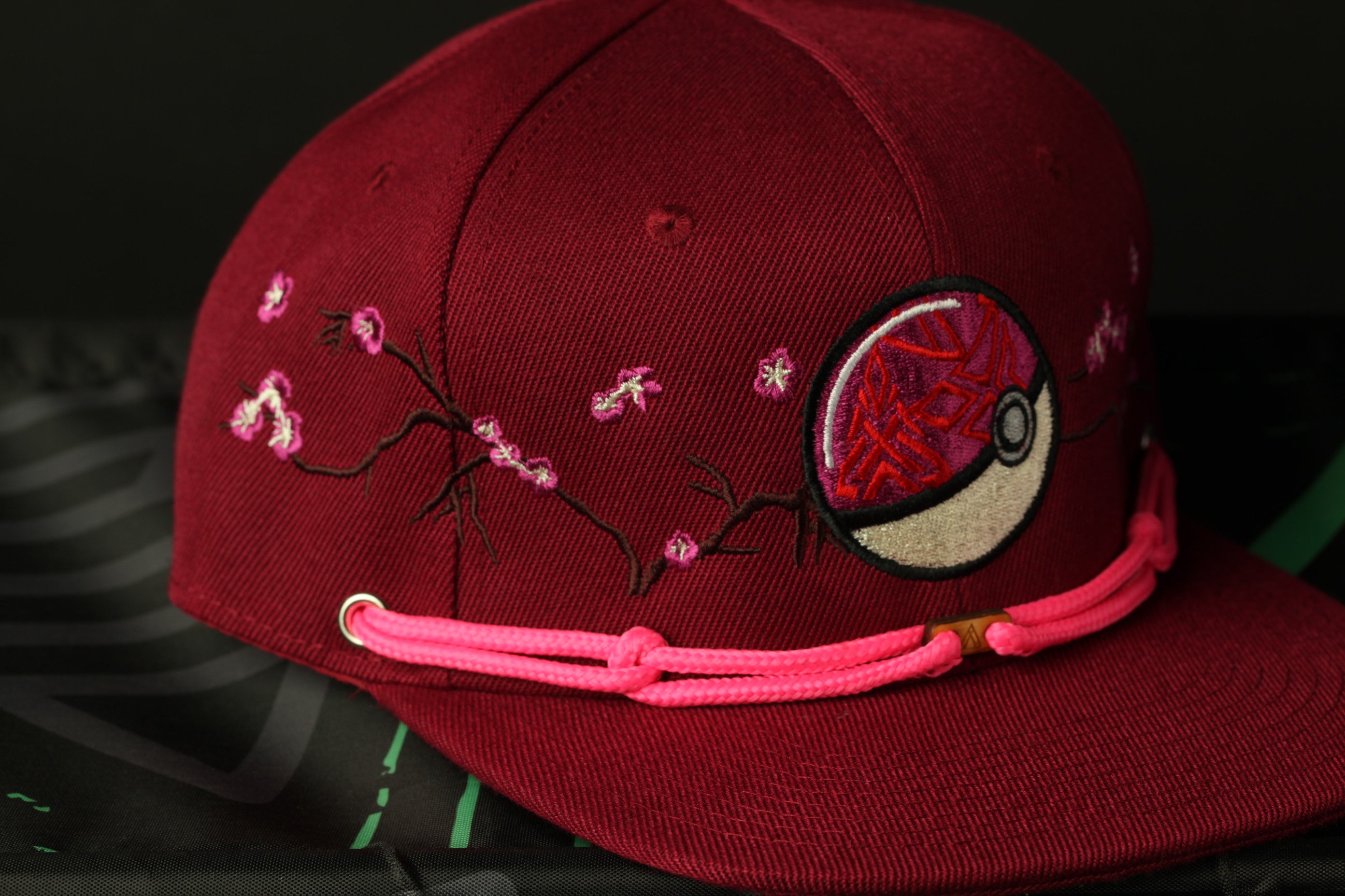 A close-up image of a maroon baseball cap with pink floral embroidery and a Pokemon-inspired design on the front.