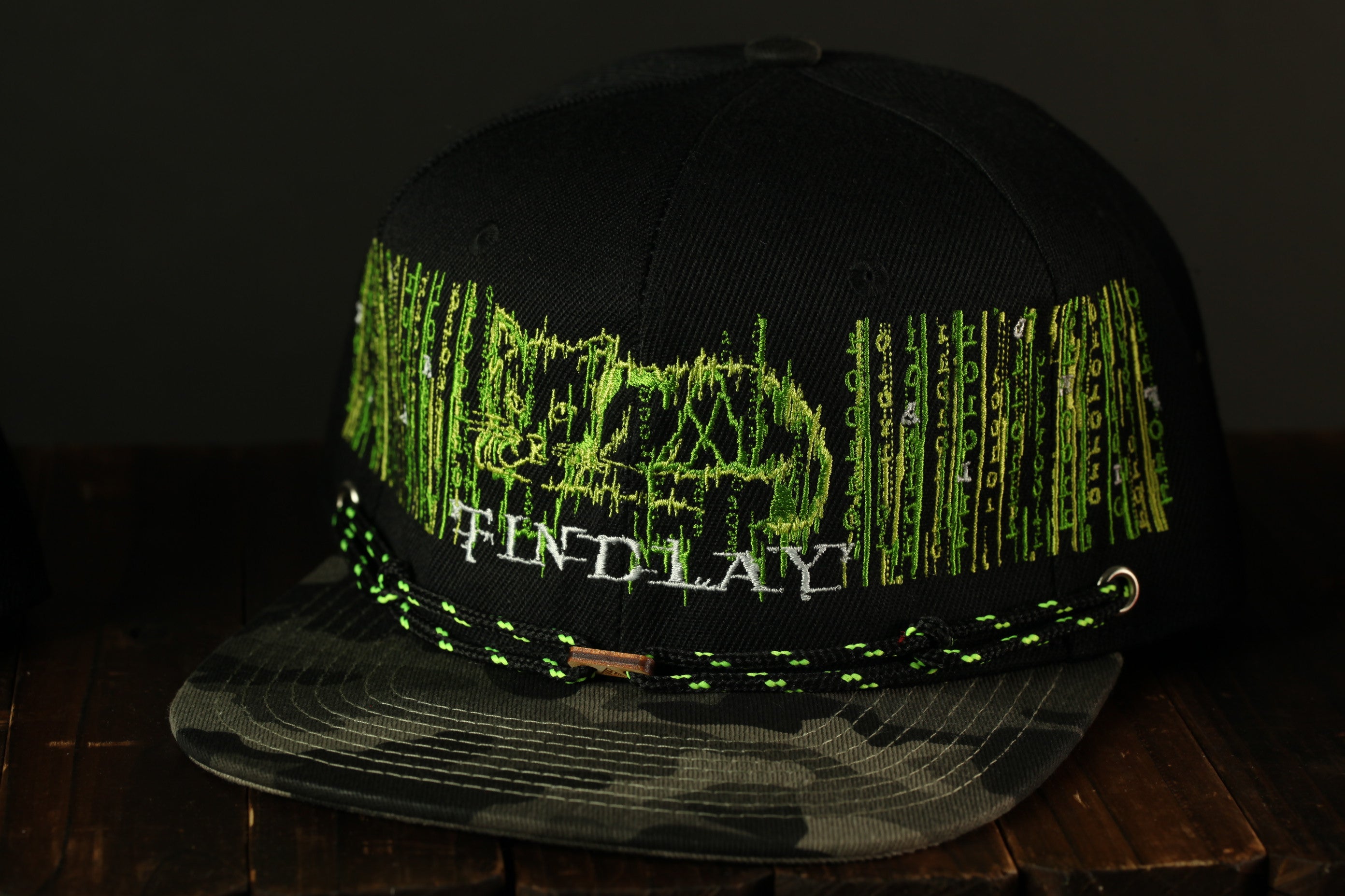 A black baseball cap with a camouflage pattern and green embroidered text on the front.