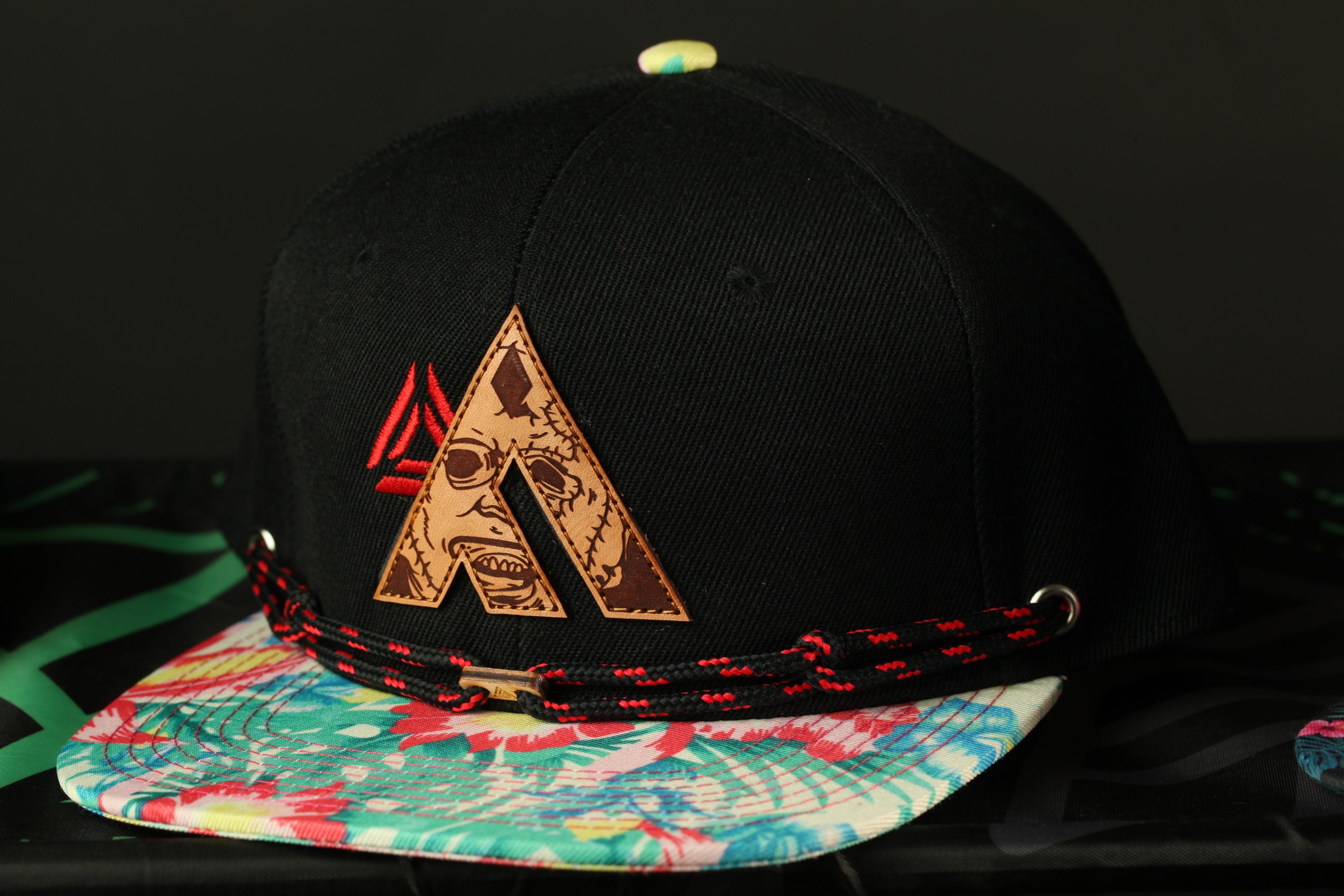 A black baseball cap with a floral pattern and a large letter "A" on it, sitting on a surface with a colorful pattern.