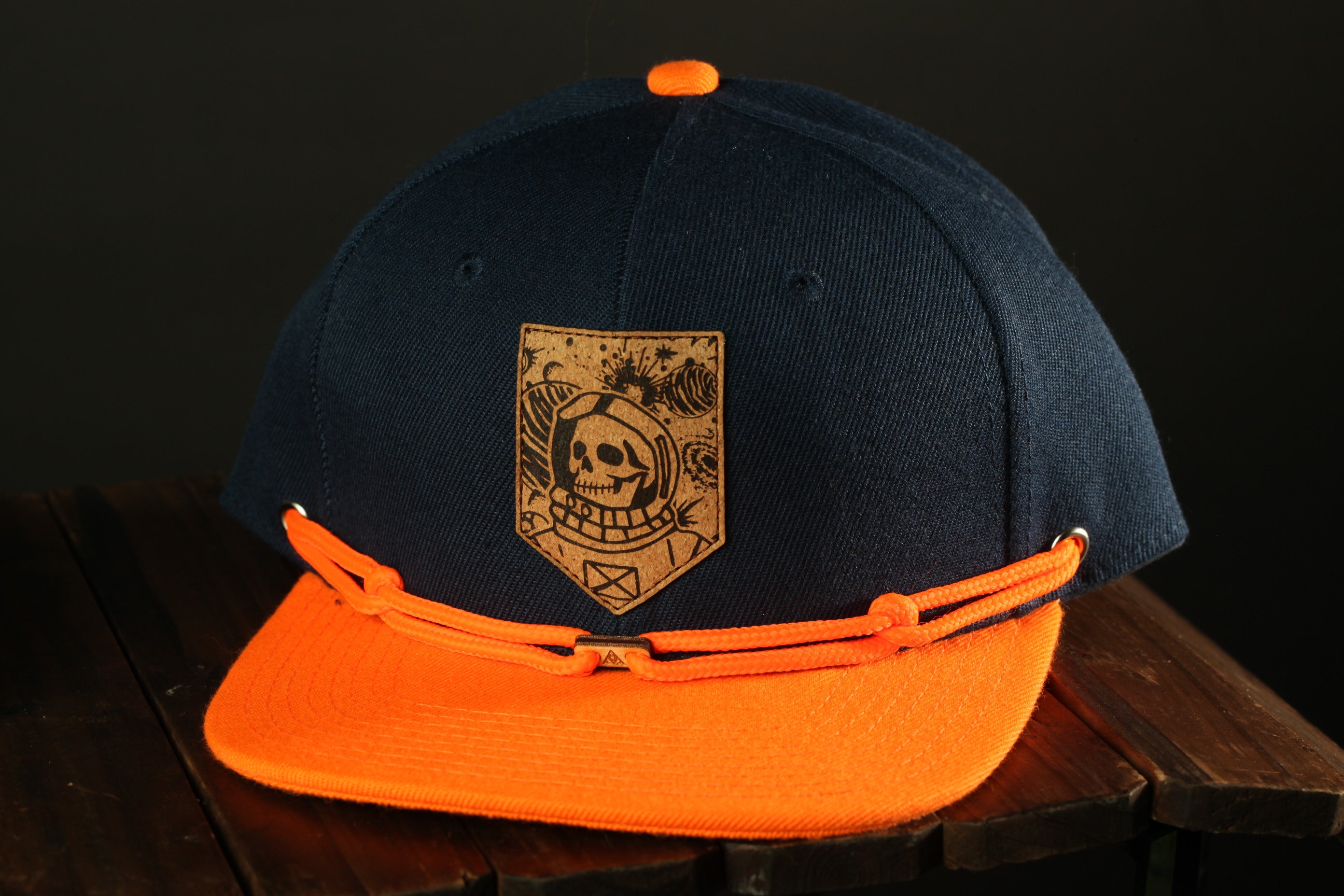 A navy blue baseball cap with an orange brim and a skull and crossbones design on the front, sitting on a wooden surface.