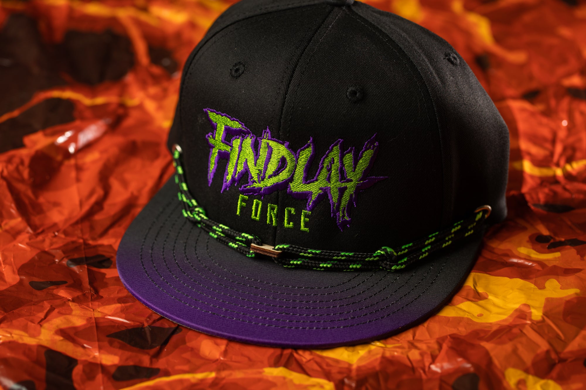 A black baseball cap with a purple brim and green and purple text on it, placed on a red and orange background.