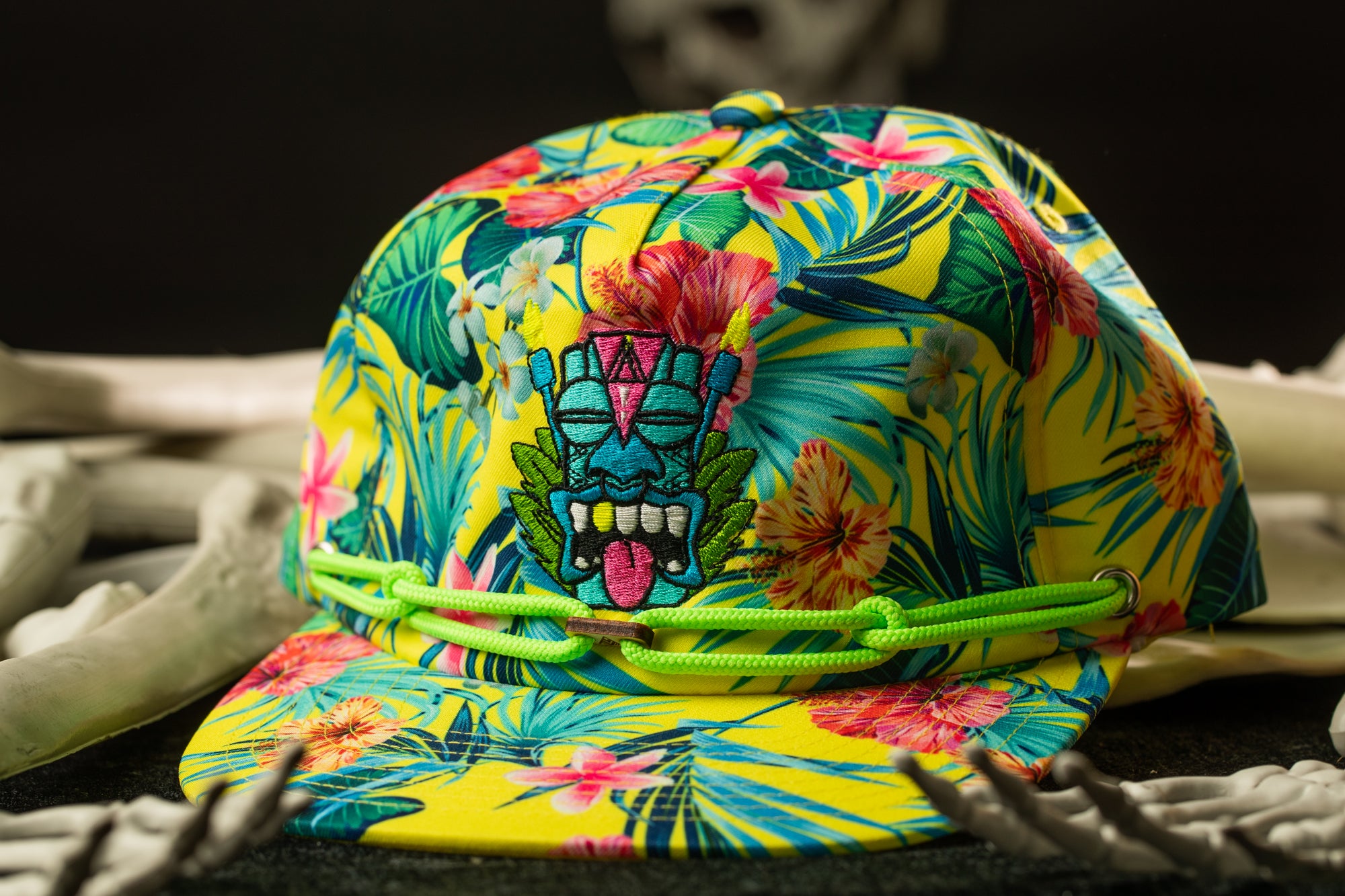 A colorful, patterned baseball cap with a tropical design, including a tiki mask and flowers, is displayed on a surface with skeletal bones.