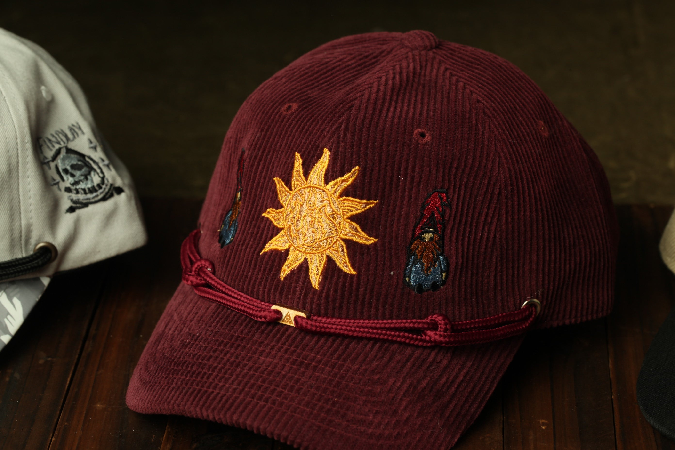 A burgundy baseball cap with a sun embroidered on it, featuring a gnome design, is displayed on a wooden surface.