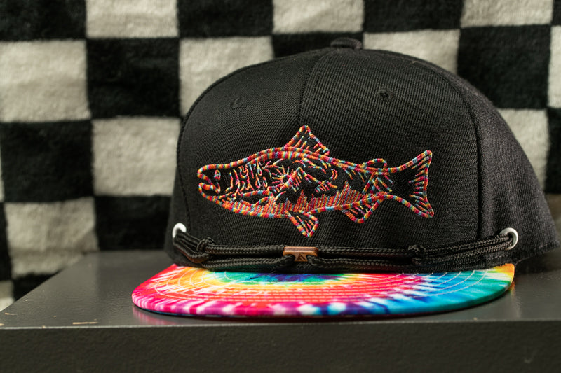 A black baseball cap with a colorful tie-dye pattern and a fish embroidered on the front, placed on a surface against a checkered background.