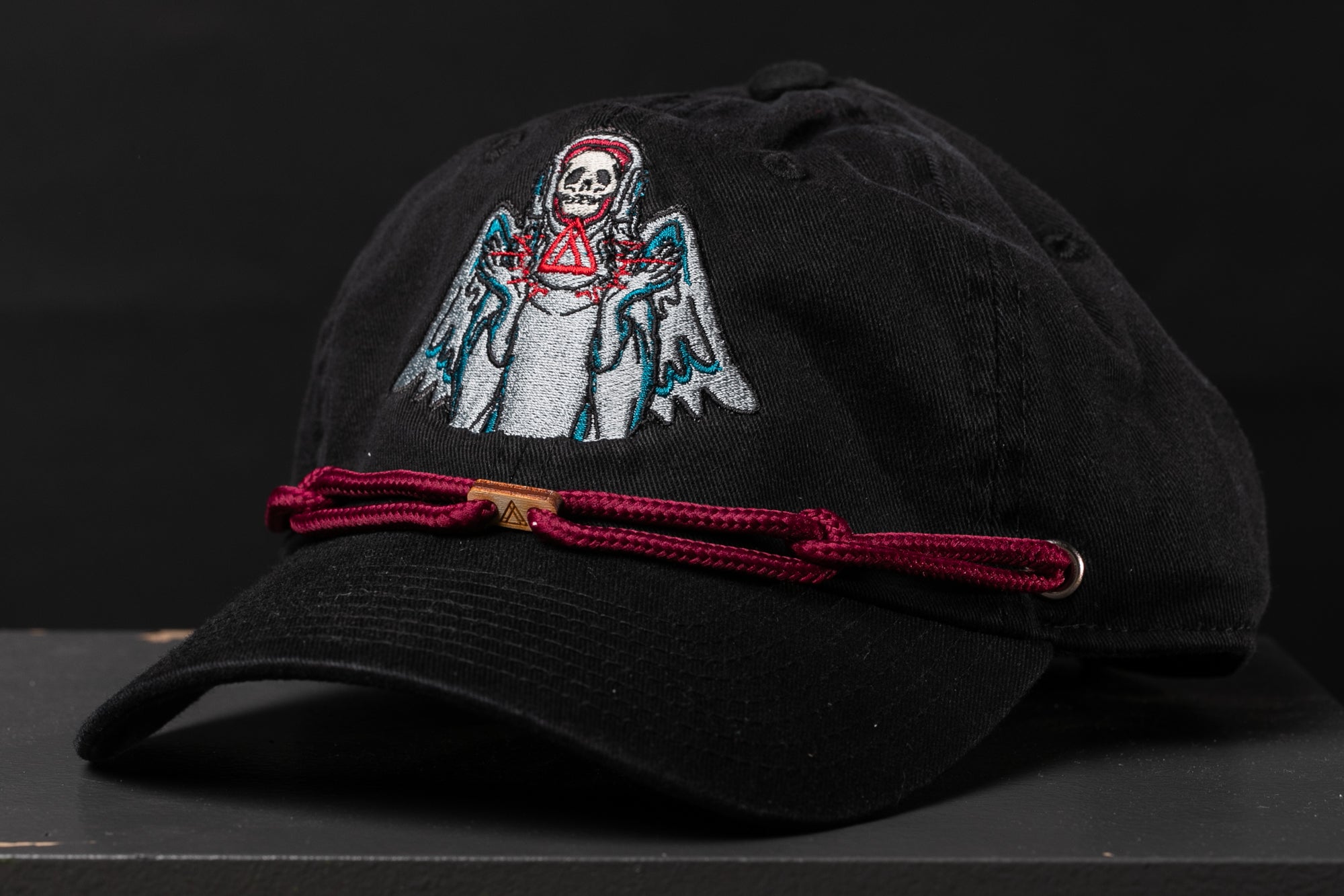A black baseball cap with a skull and crossbones design on the front, and a red string or strap around the brim.
