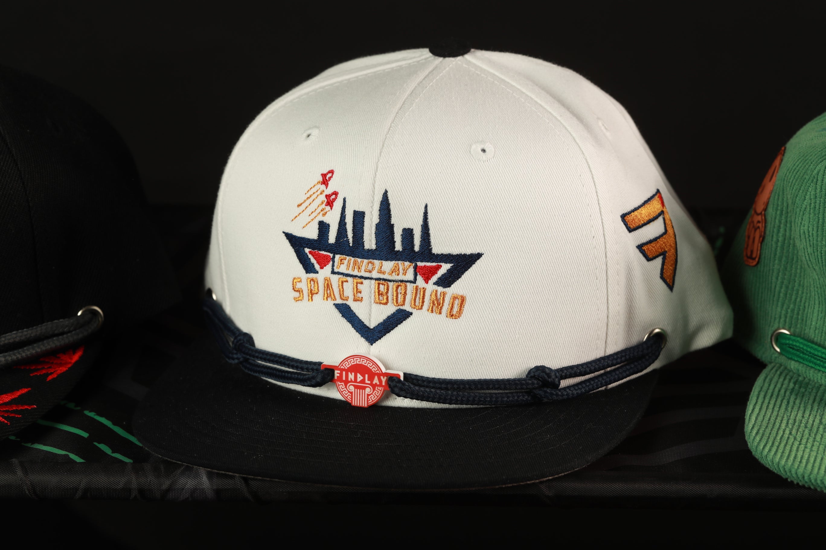 A white baseball cap with a black brim and a red "FINOLAY SPACE BOUND" logo, along with a black and red lanyard, is displayed among other hats.