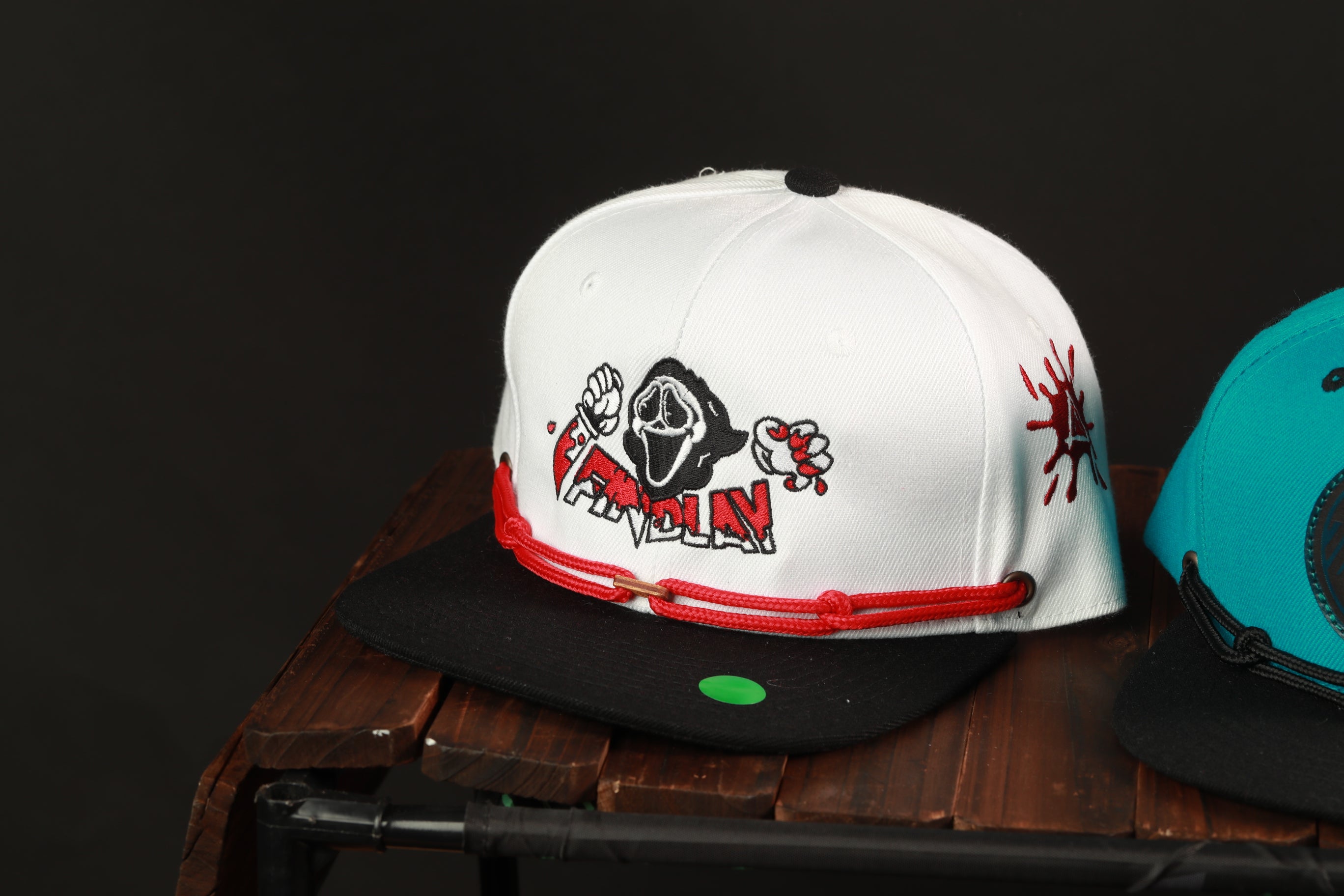 A white baseball cap with a graphic design featuring a hooded figure and the word "SHOOTER" printed on it, sitting on a wooden surface.