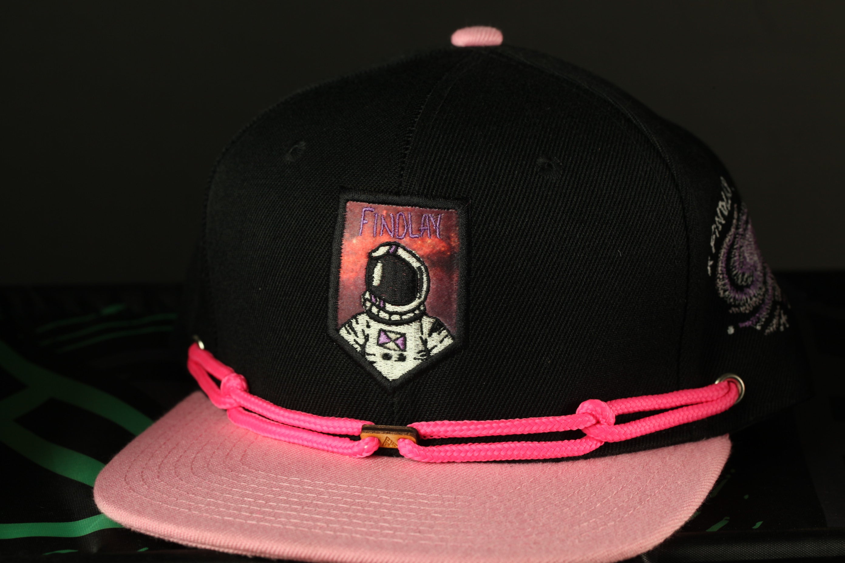 A black baseball cap with a pink strap and a patch featuring an astronaut.