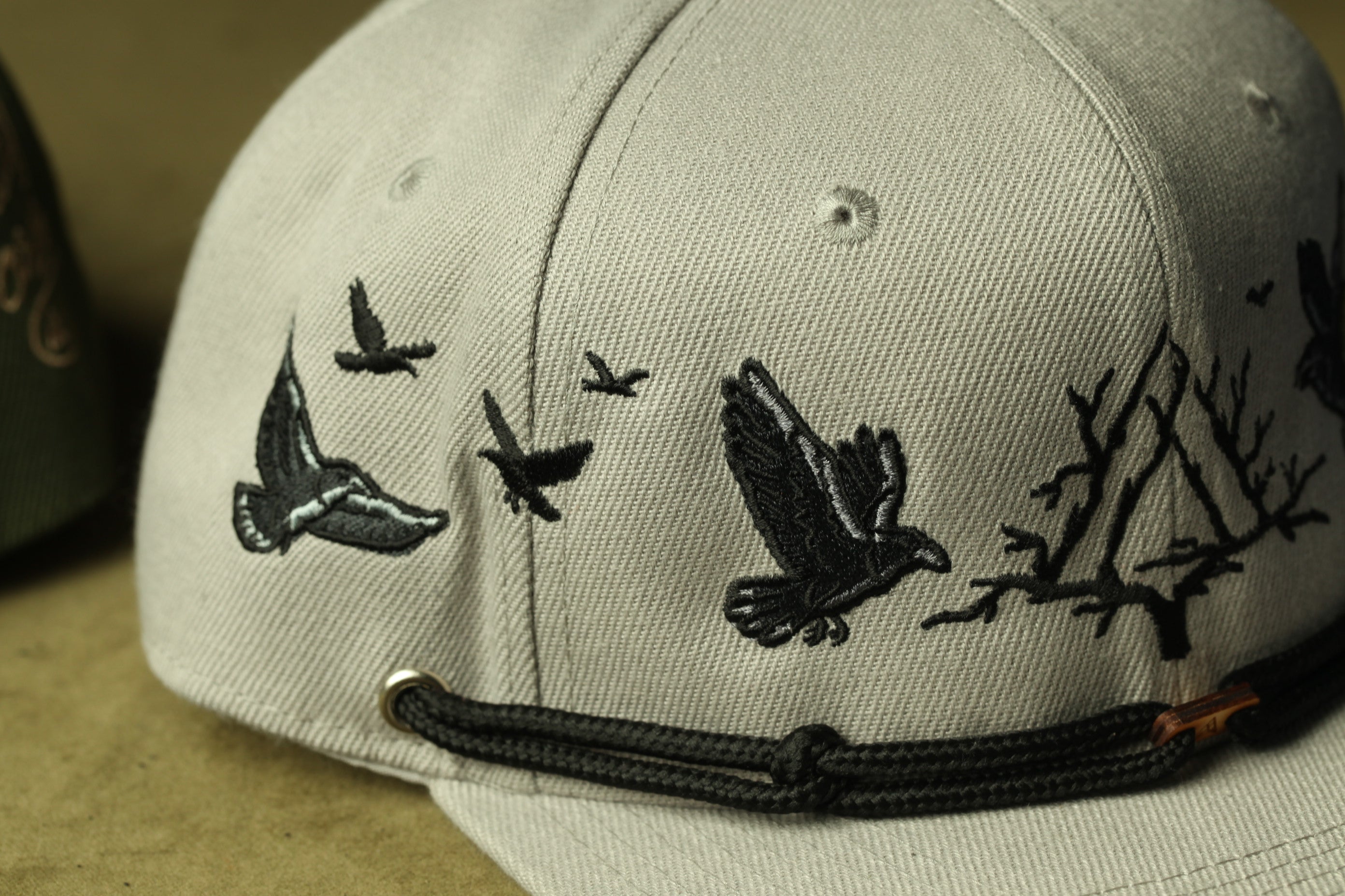 A close-up view of a gray baseball cap with black bird embroidery, featuring silhouettes of birds in flight.