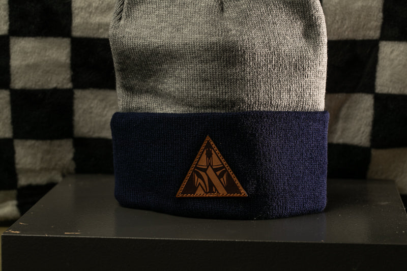 A gray and navy blue beanie with a brown triangular patch on the front, placed on a black surface against a checkered background.