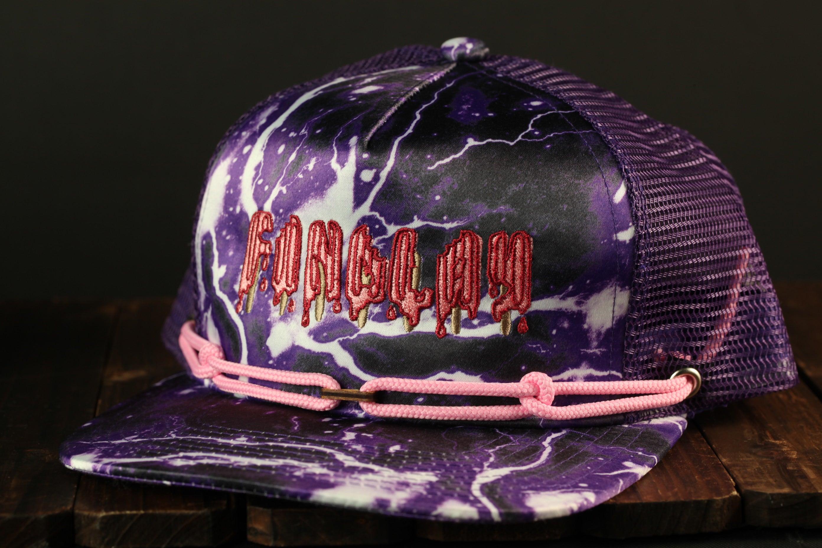 A purple and white baseball cap with the word "Fungus" printed on it, sitting on a wooden surface.