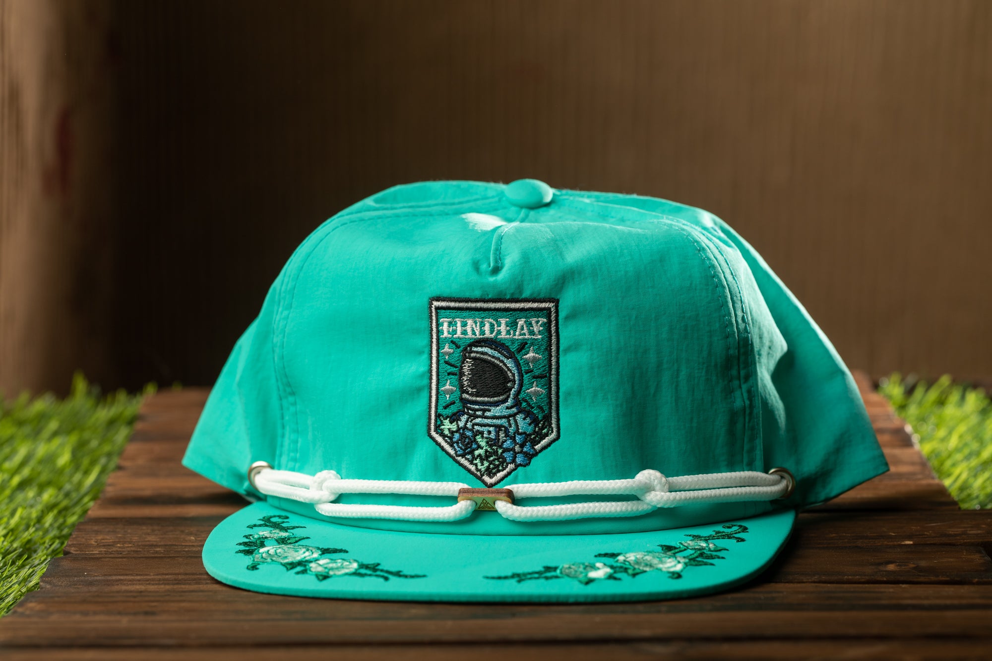 A turquoise baseball cap with a patch featuring a space helmet and the word "INDELAY" printed on it, placed on a wooden surface.