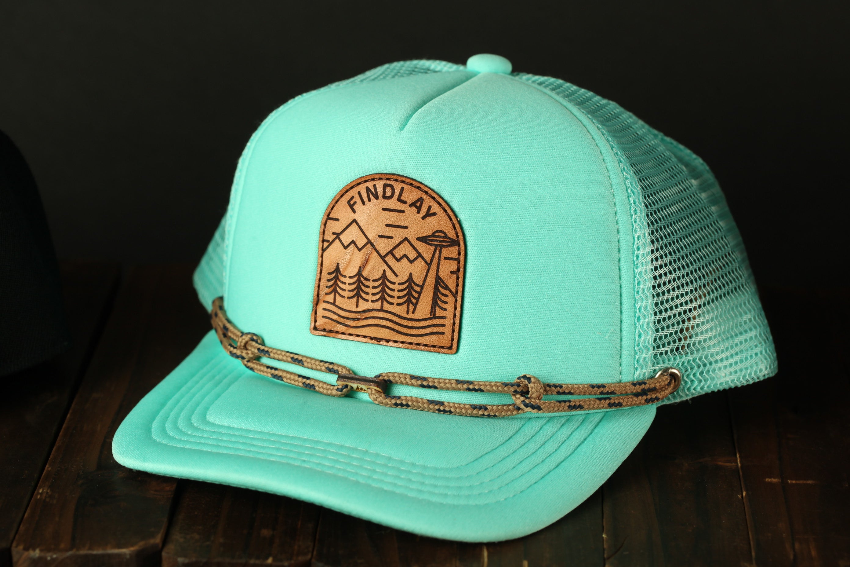A turquoise baseball cap with a leather patch on the front, featuring a mountain landscape design.