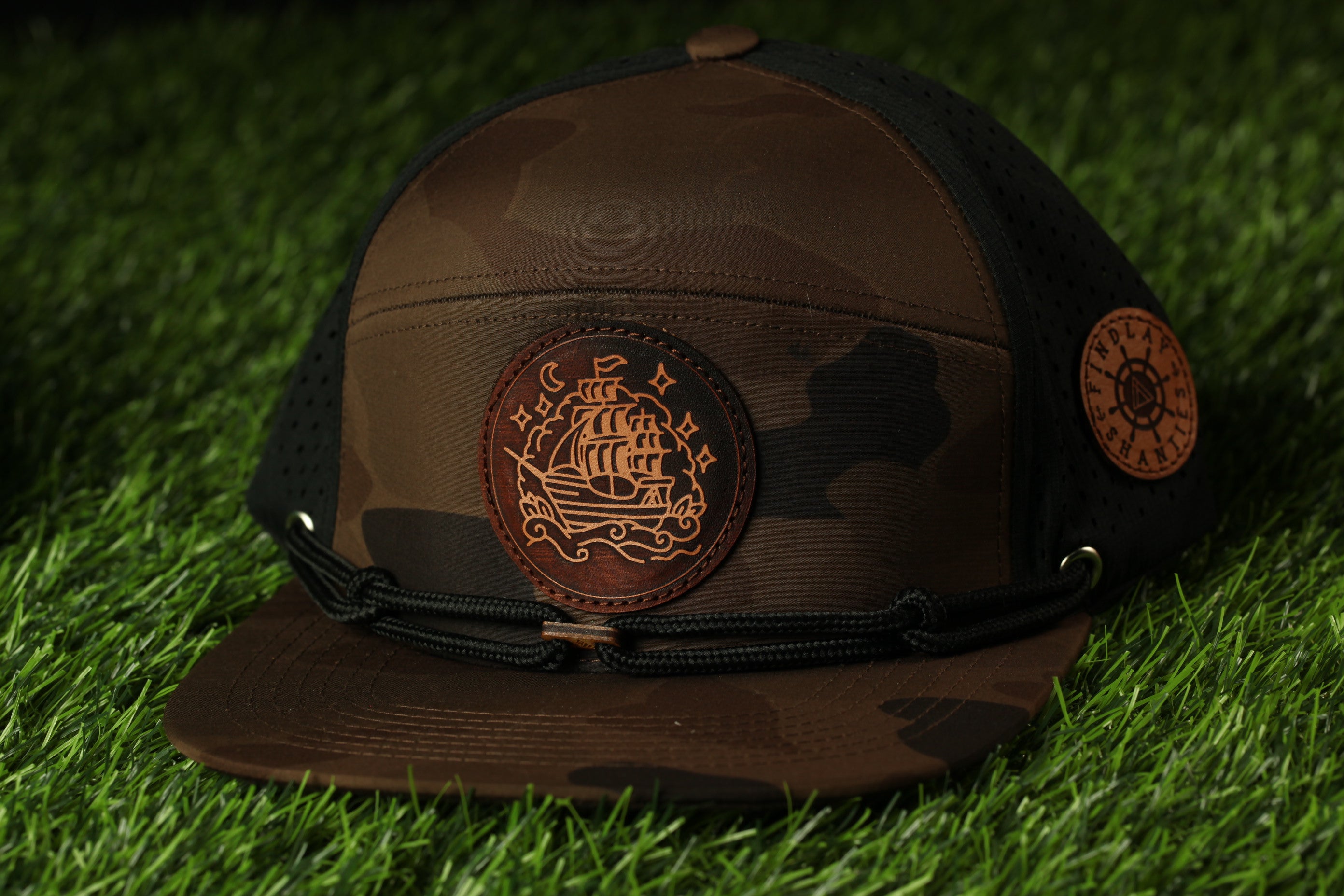 A brown camouflage baseball cap with a ship embroidered on it, placed on a grassy surface.