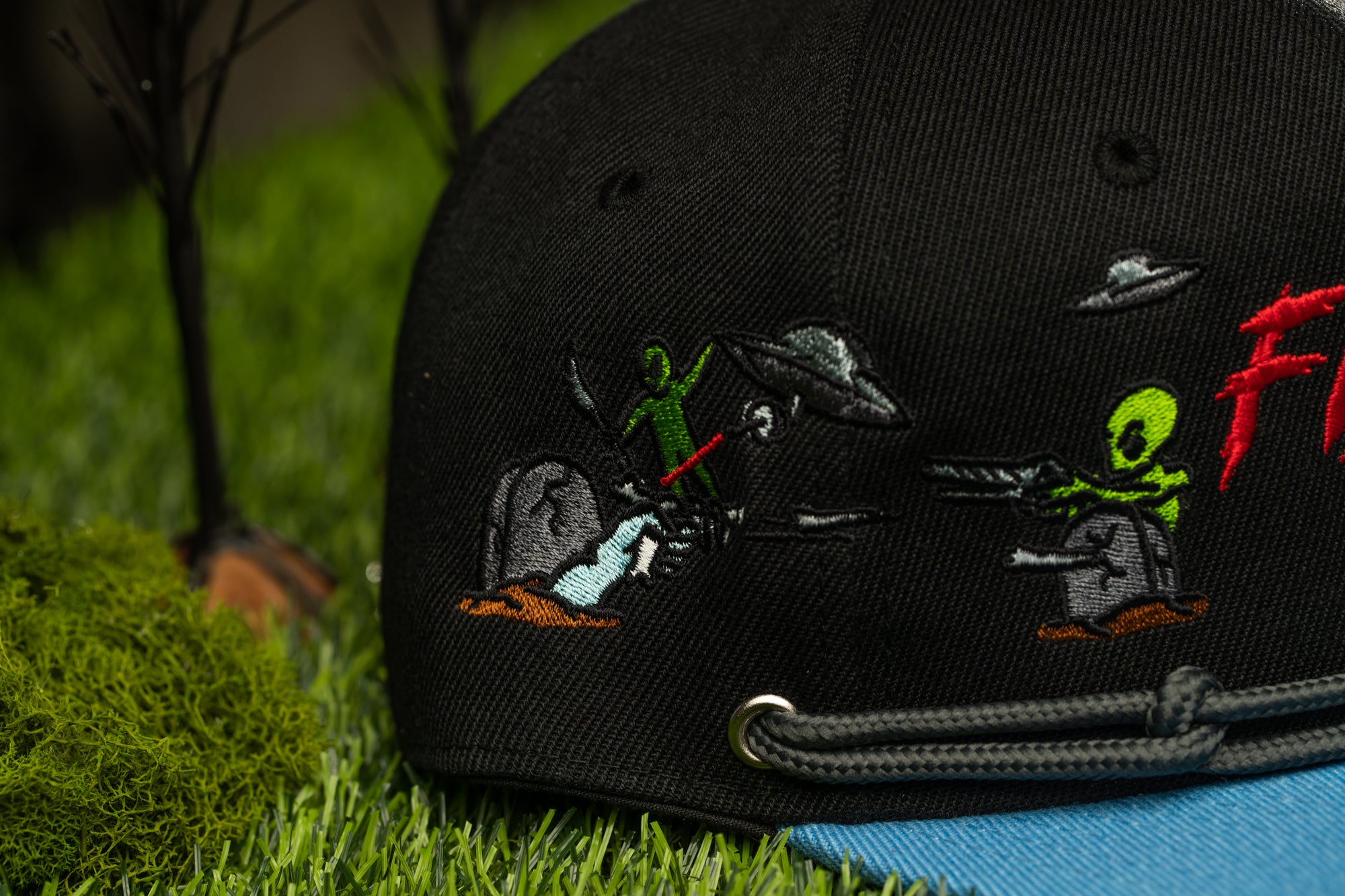 a black baseball cap with a detailed embroidered design depicting a scene with green alien characters and a spaceship.